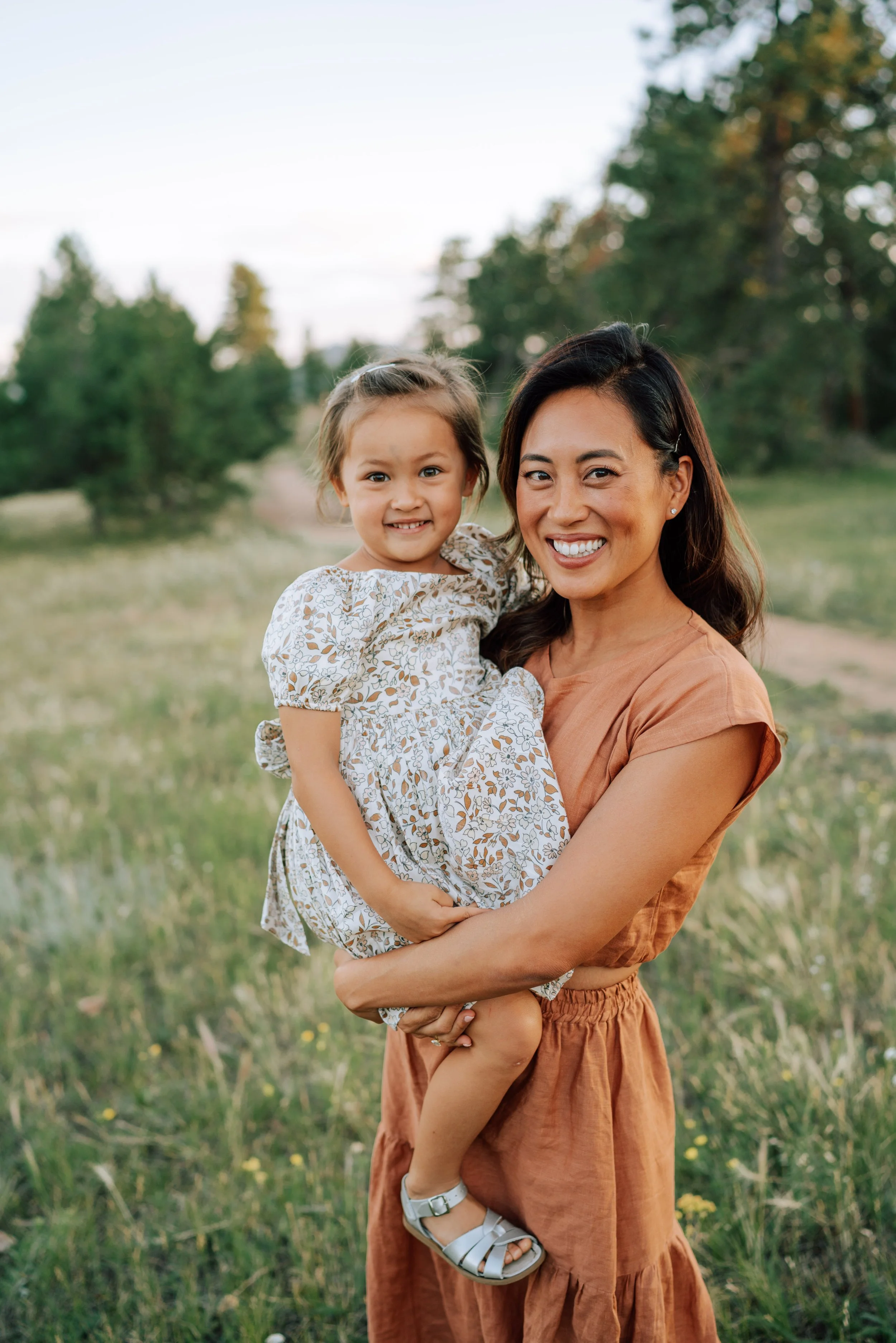 Mom holding her young daughter during an outdoor family session surrounded by soft grass and evergreen trees at Mount Falcon.
