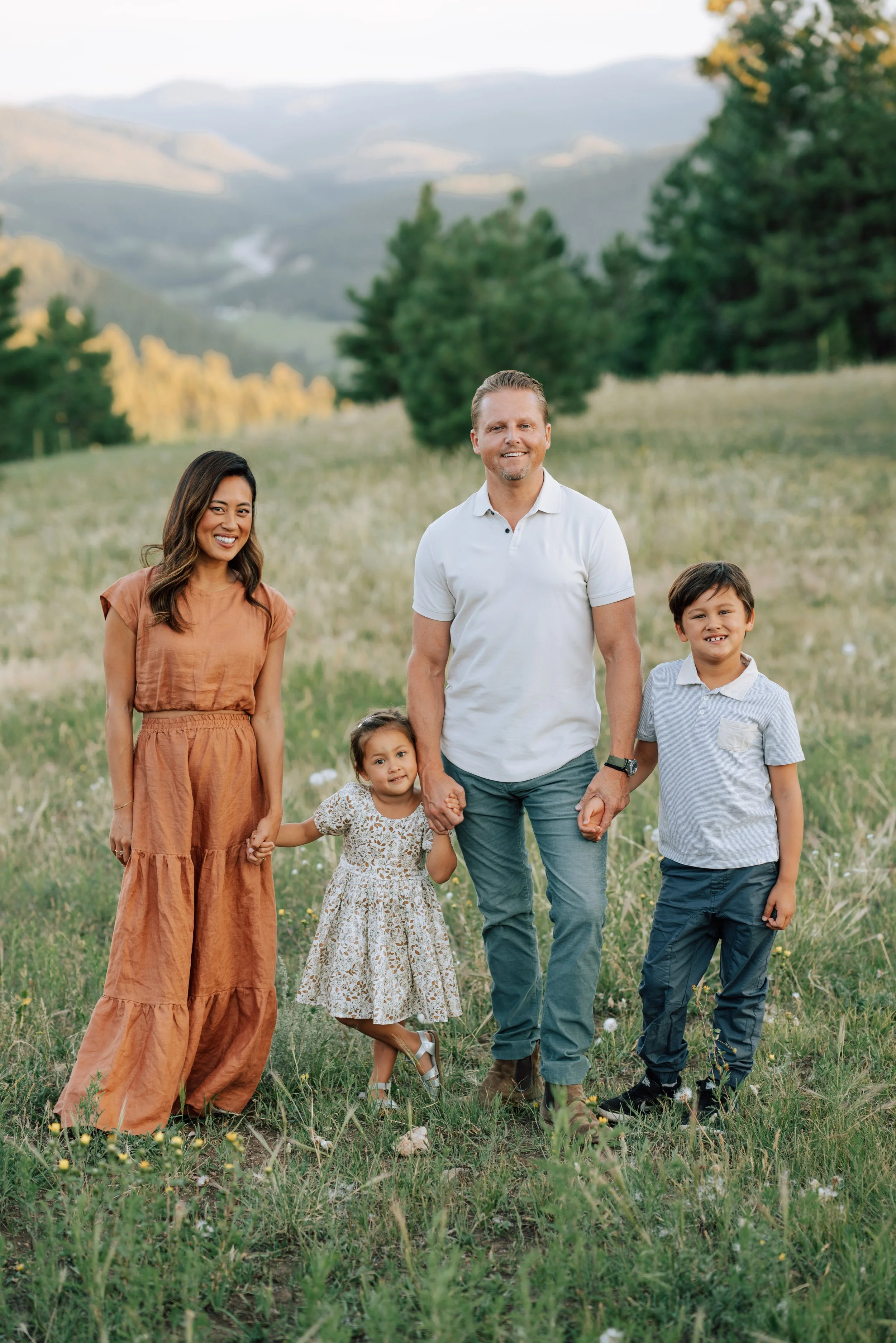 Family of four surrounded by tall summer grass with pine trees and Colorado mountains in the background at Mount Falcon.