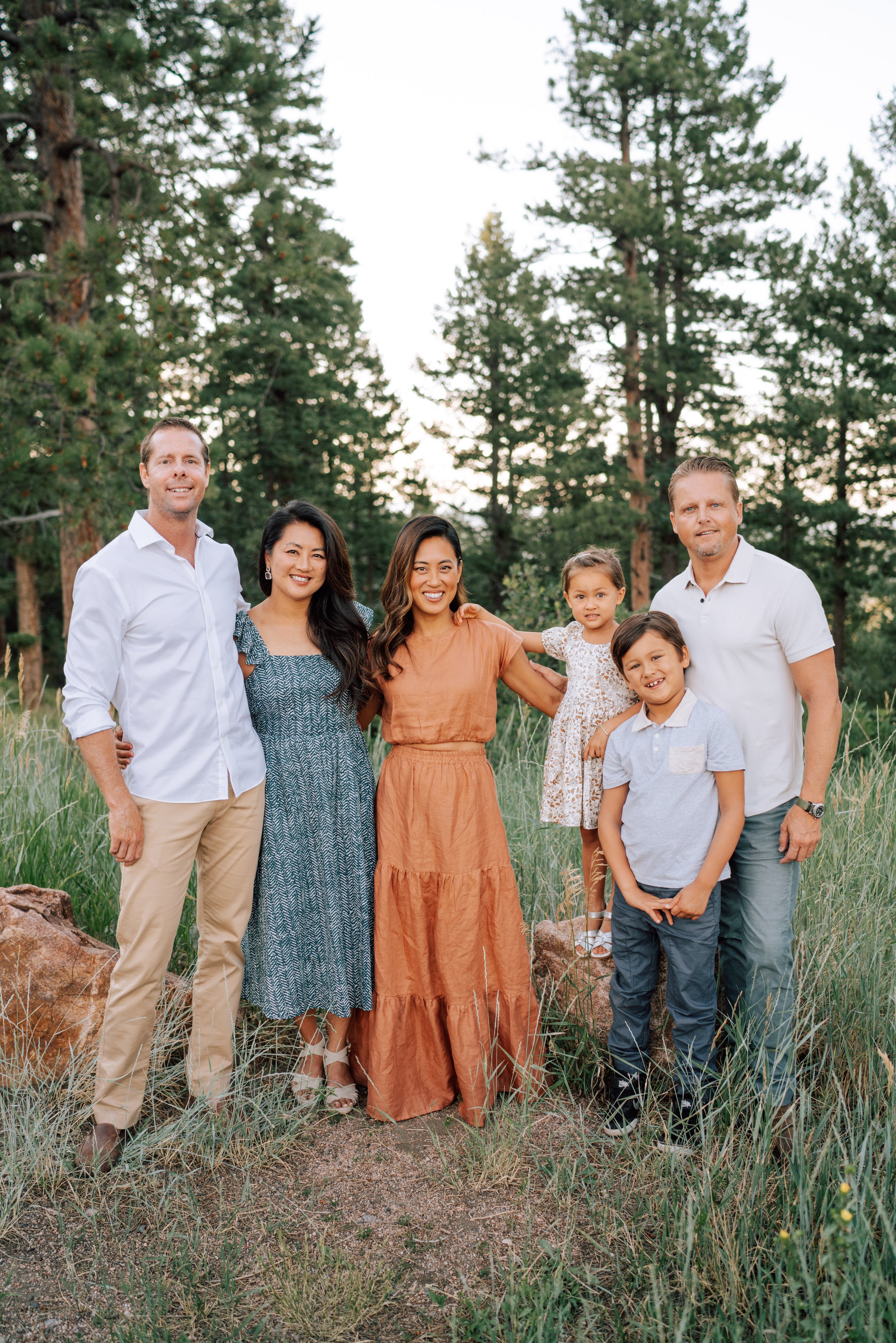 Vertical extended family portrait at Mount Falcon with two sisters, their families, tall grass, rocks, and evergreen trees.