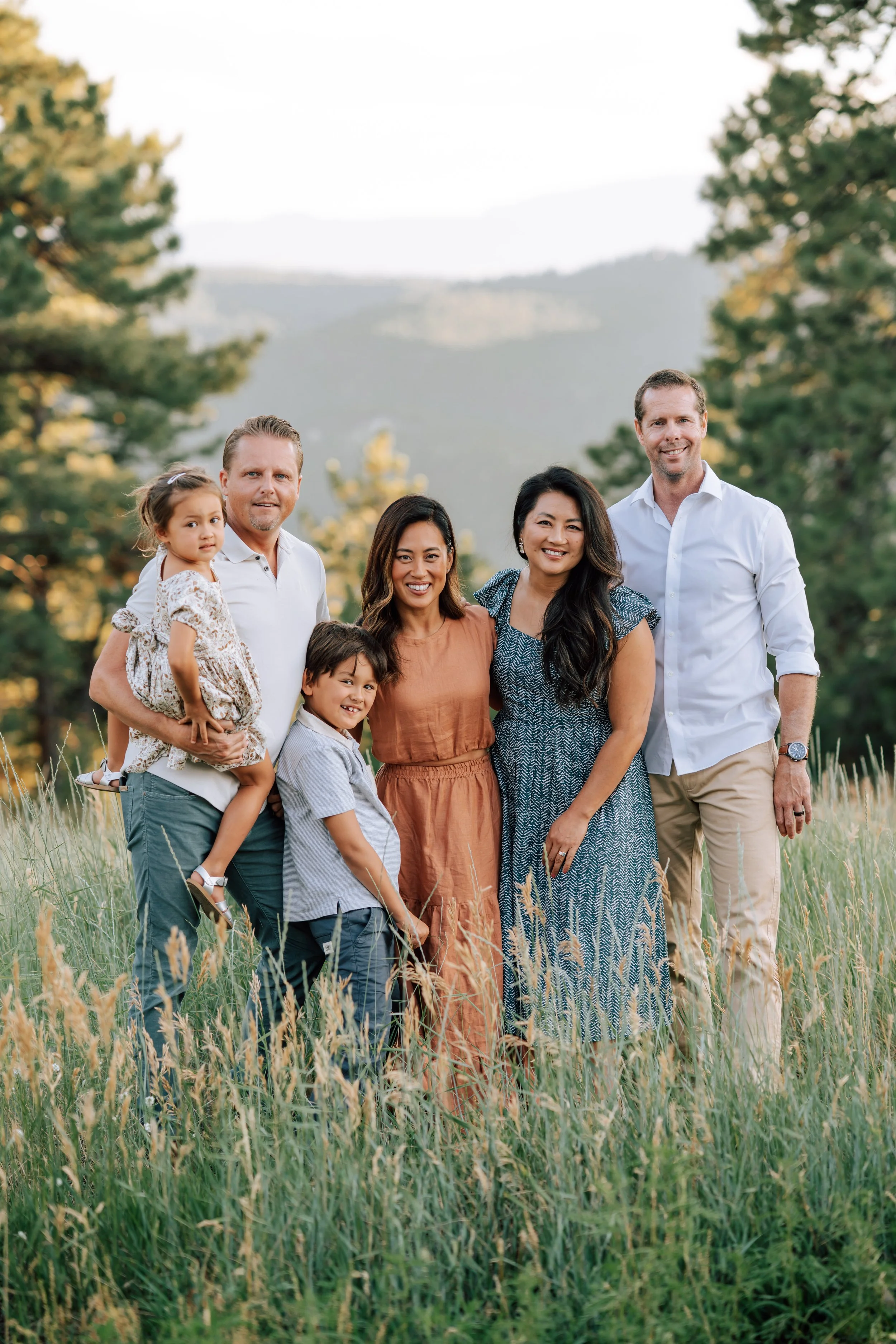 Extended family standing together in tall grass with pine trees and distant Front Range mountains during an evening family session.