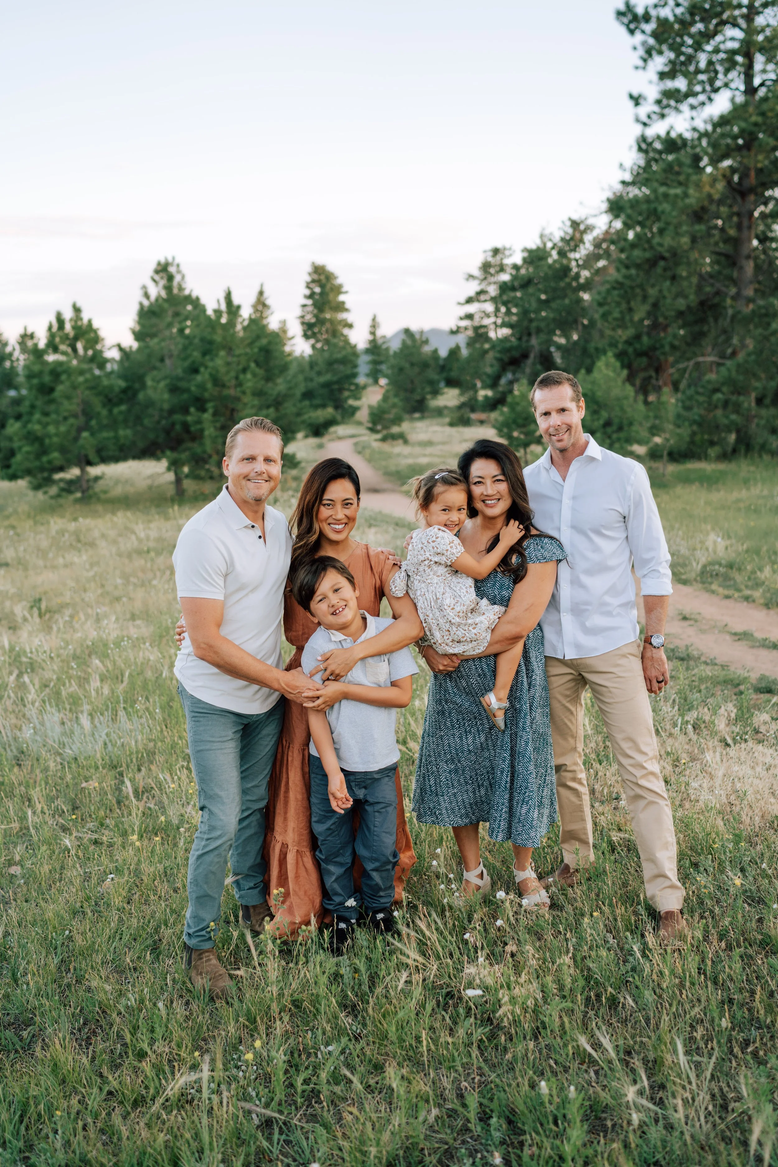 Two sisters and their families photographed together in an open meadow with children cuddled close and mountain scenery behind them.