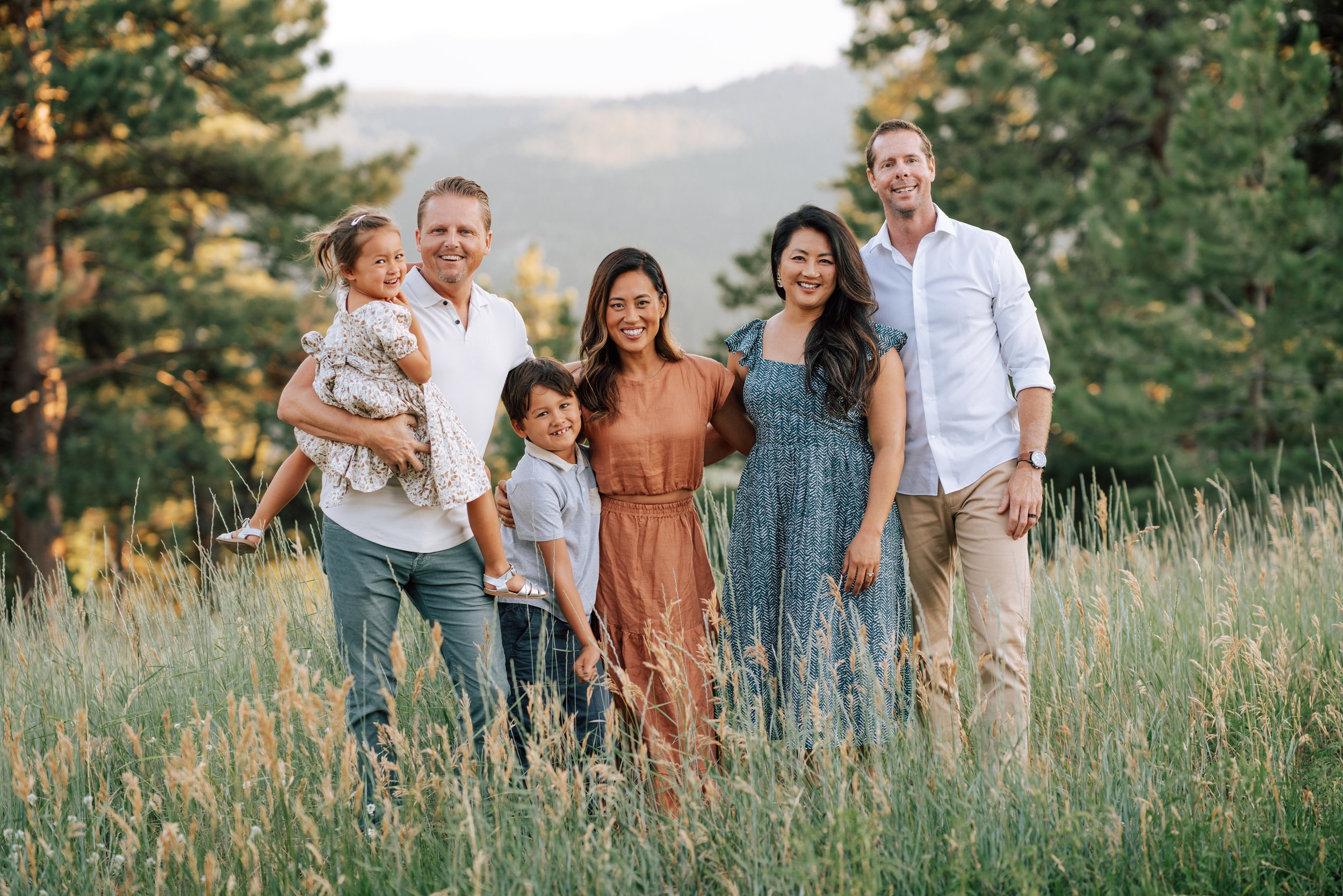 Extended family portrait in tall grass with evergreen trees and layered Colorado mountain views at Mount Falcon West.