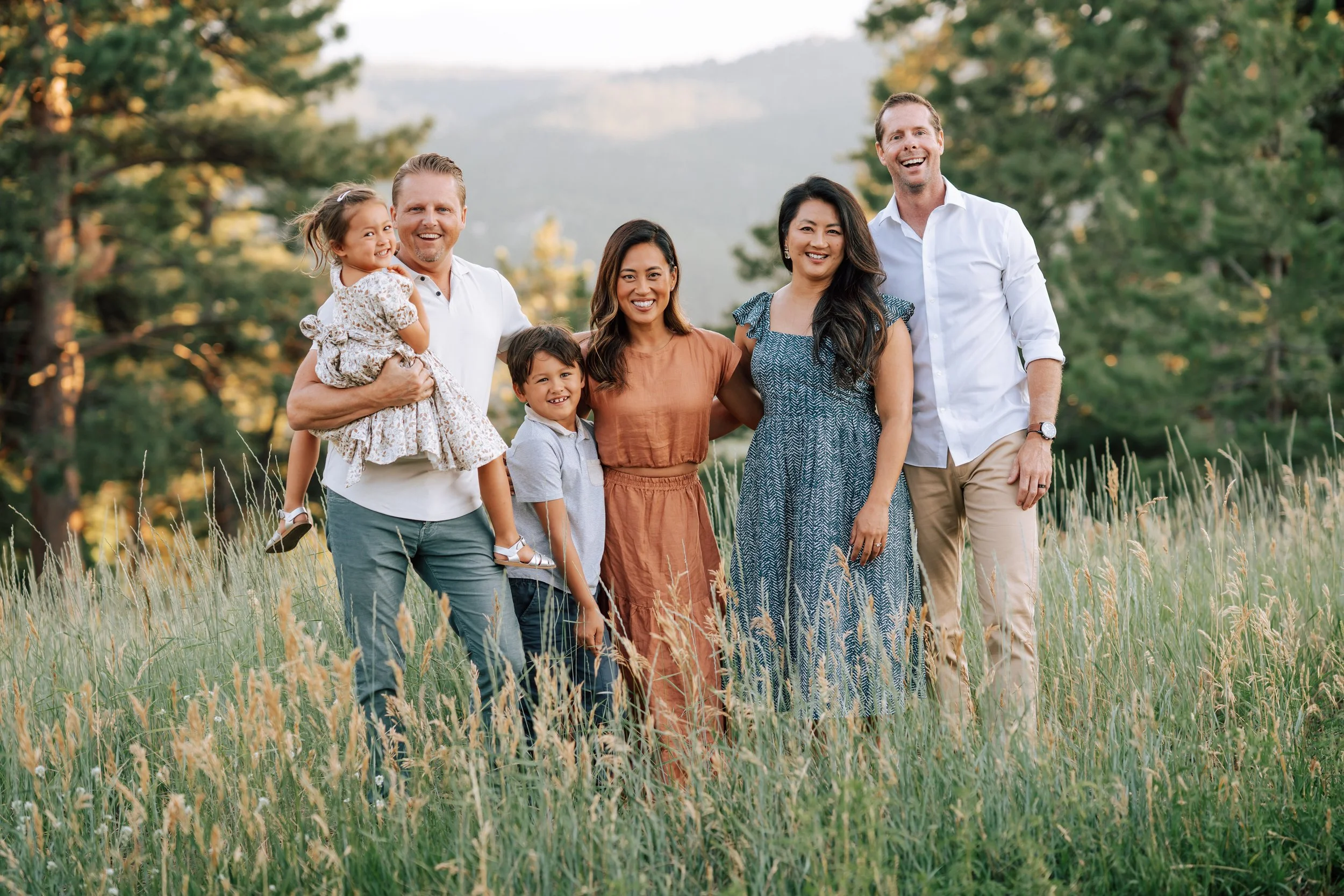 Extended family portrait in tall grass with evergreen trees and layered Colorado mountain views at Mount Falcon West.