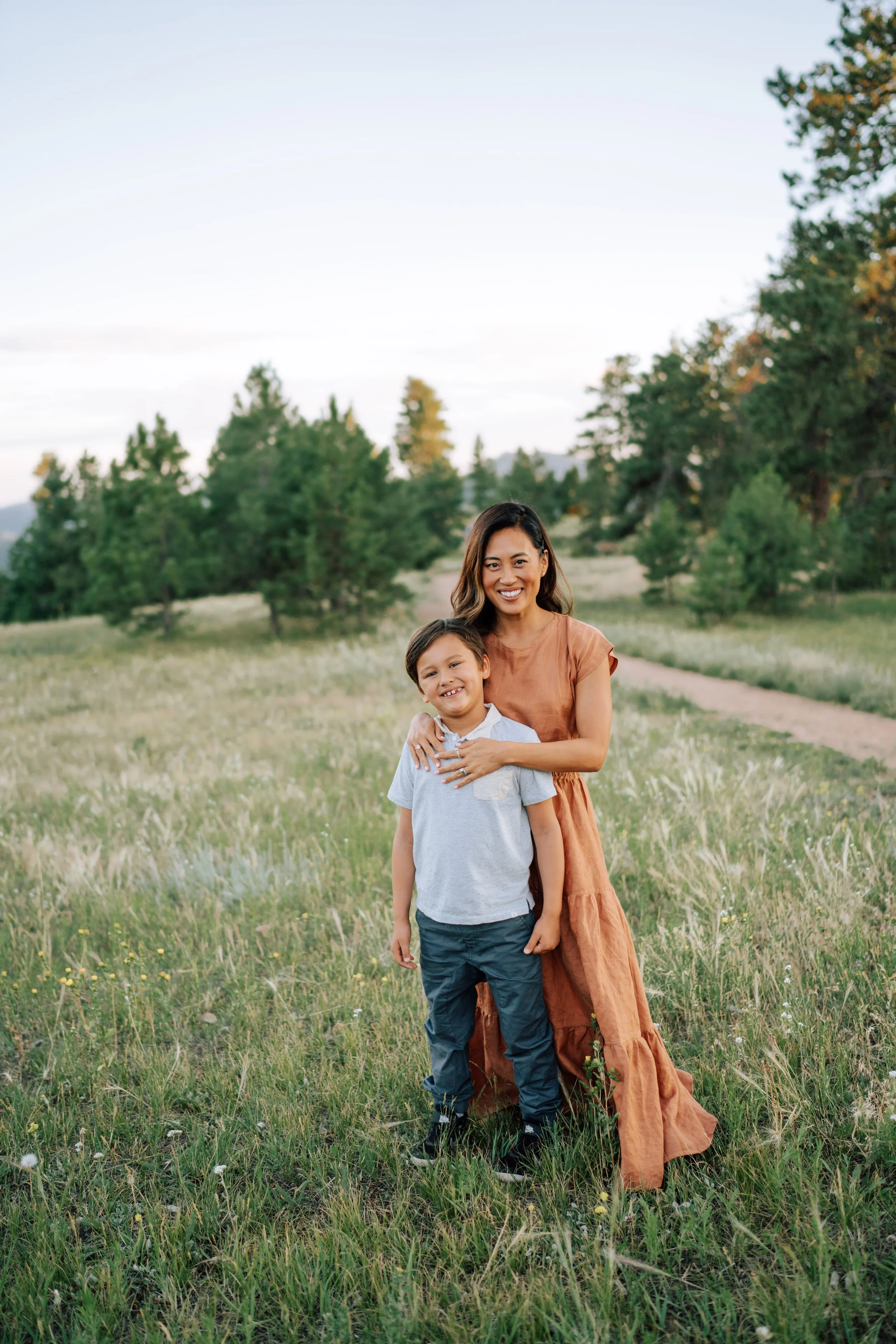 Mom standing with her young son in a grassy Mount Falcon meadow with pine trees and a trail in the background.