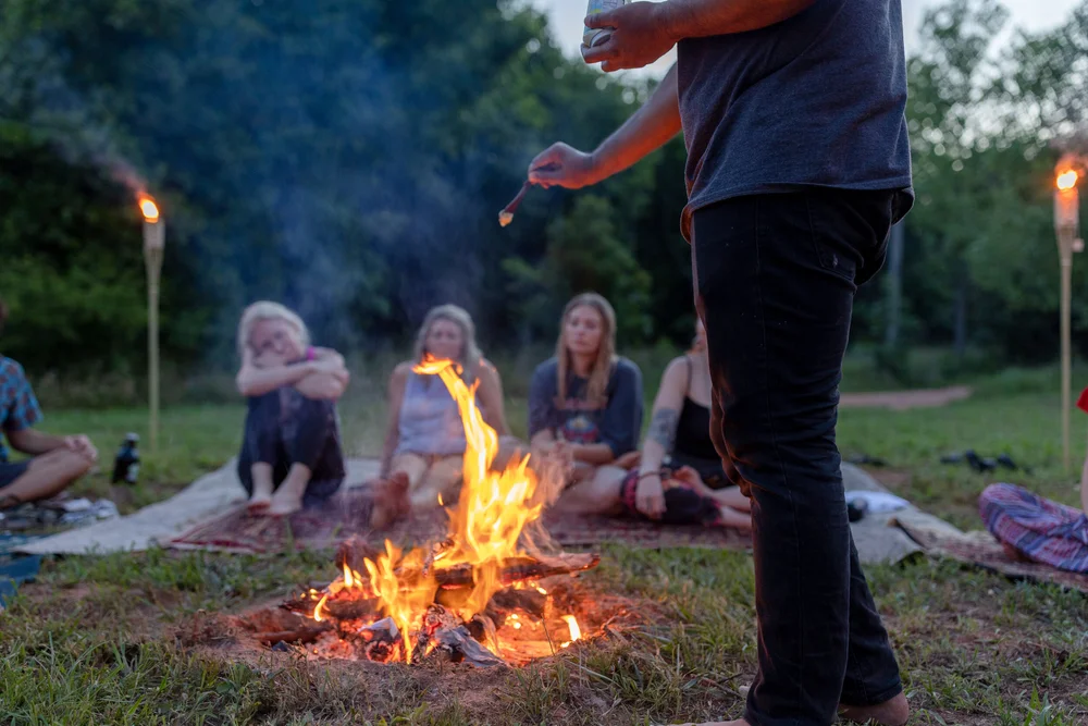 A man standing by a campfire at dusk as a group sits around it on blankets