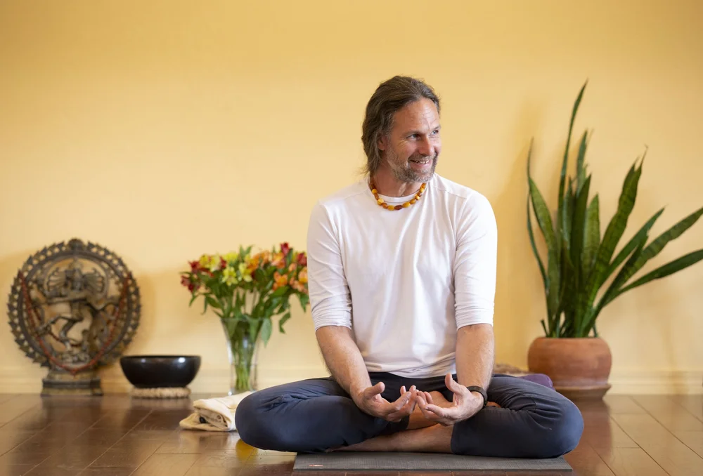 A man sitting in lotus and smiling, beside flowers and a brass gong