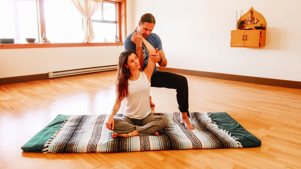 A seated Thai bodywork stretch on a striped mat, the arms drawn back