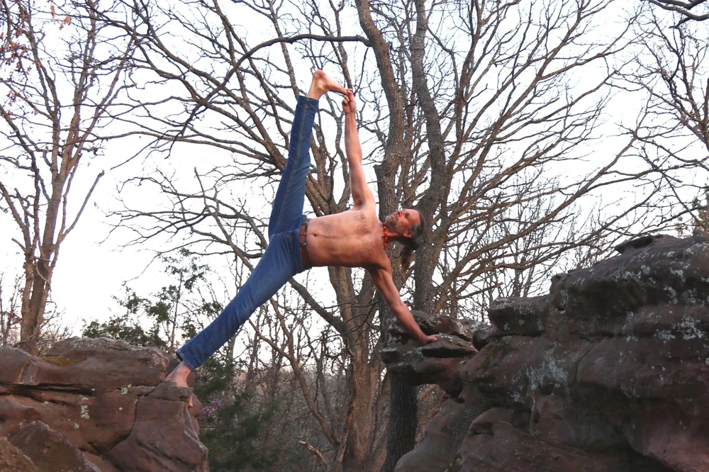 A yogi balancing in a standing split on a sandstone outcrop against bare winter trees