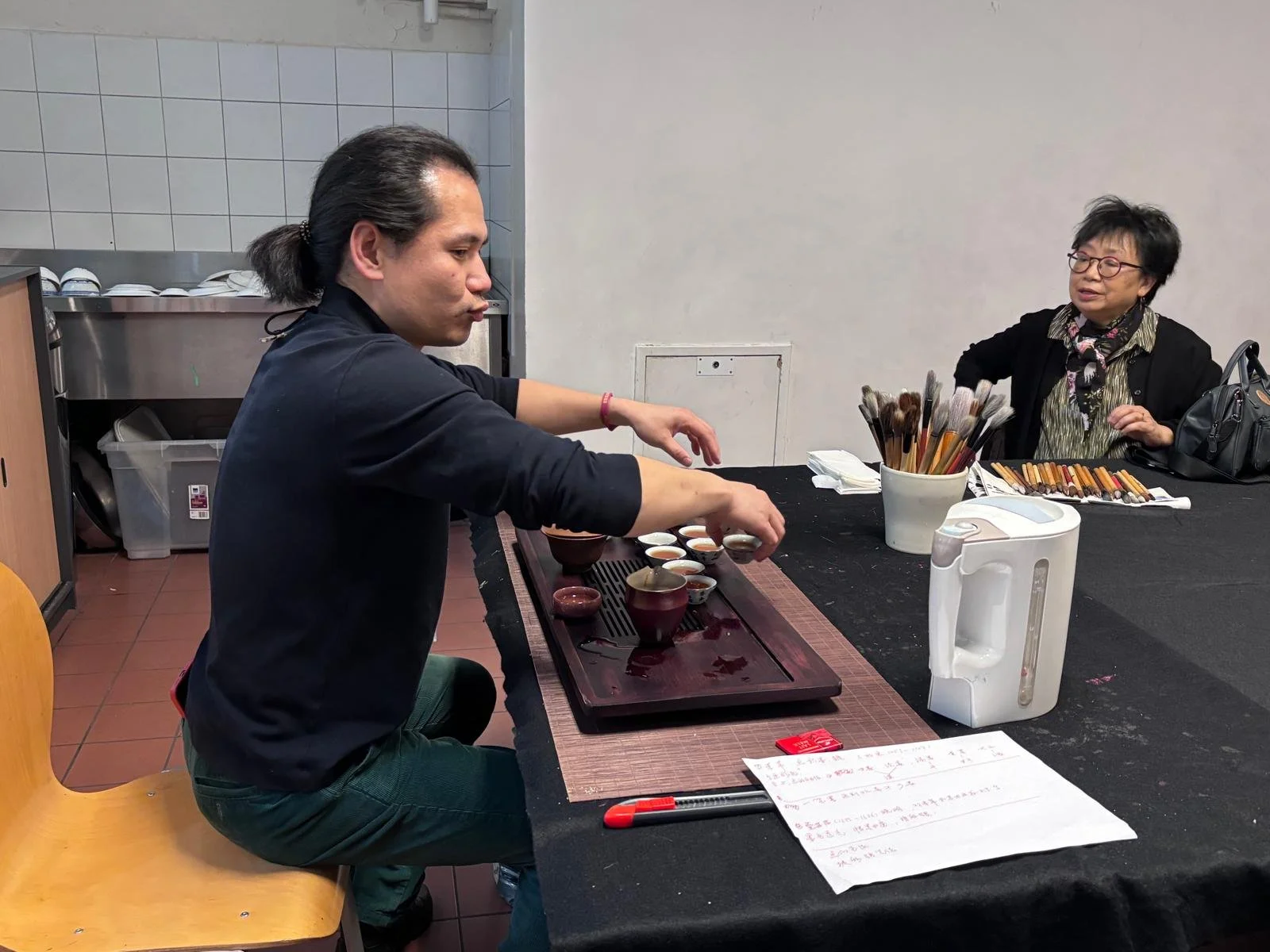 Un homme prépare du thé ou une boisson chaude dans un établissement, avec une femme assise en face, entourée de pinceaux de peinture et d'outils artistiques sur la table.