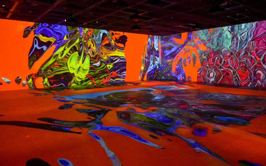 Une salle d'exposition avec des projections de motifs colorés et abstraits sur les murs et le sol, créant une ambiance immersive.