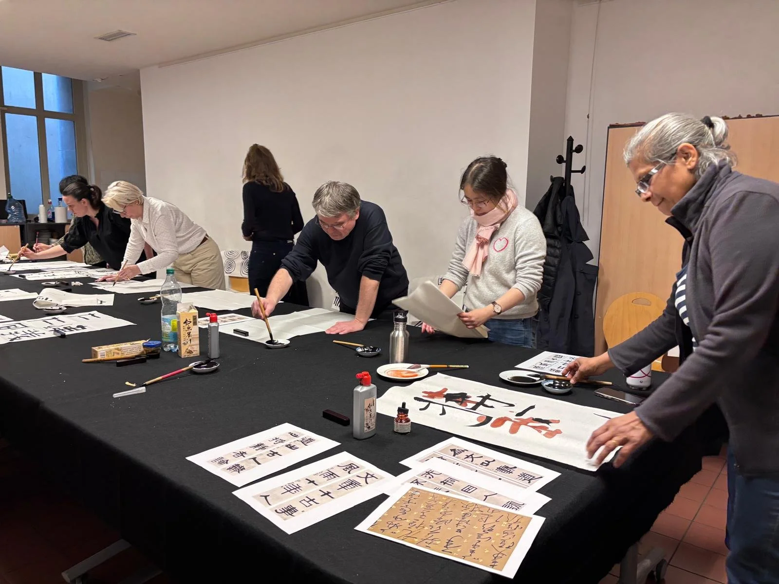 Groupe de personnes pratiquant la calligraphie japonaise sur une grande table, avec des pinceaux, encre, et papiers calligraphiques, dans une salle éclairée par une lumière naturelle.
