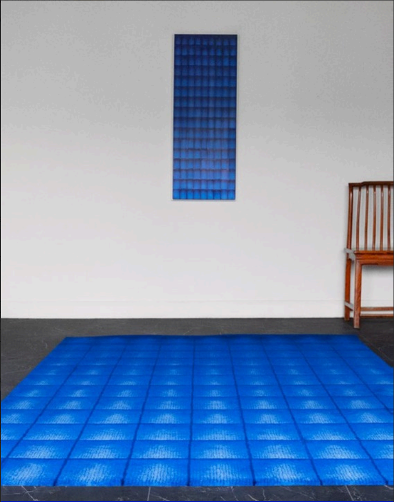 Une pièce avec un tapis bleu en mosaïque au sol, une pièce murale blanche avec une sculpture murale rectangulaire bleue, et un banc en bois à droite.