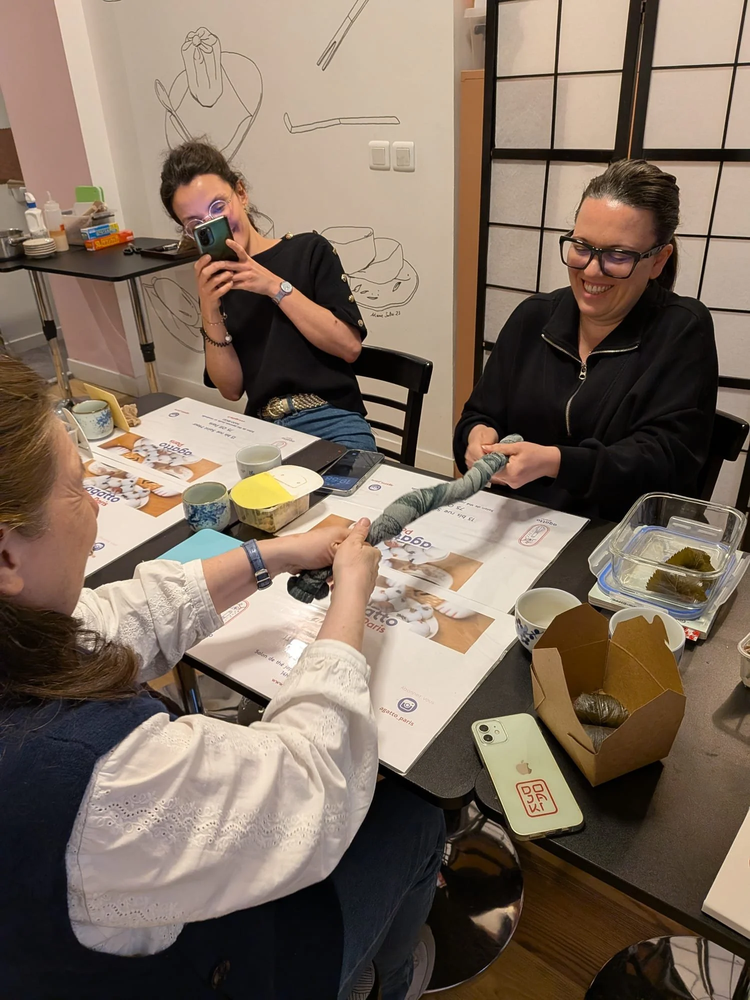 Trois femmes rient et s'amusent autour d'une table, participant à une activité de nouage avec une corde ou un fil. La table est pleine de plats, tasses, et objets divers, dont un téléphone portable et une boîte en carton avec des aliments.