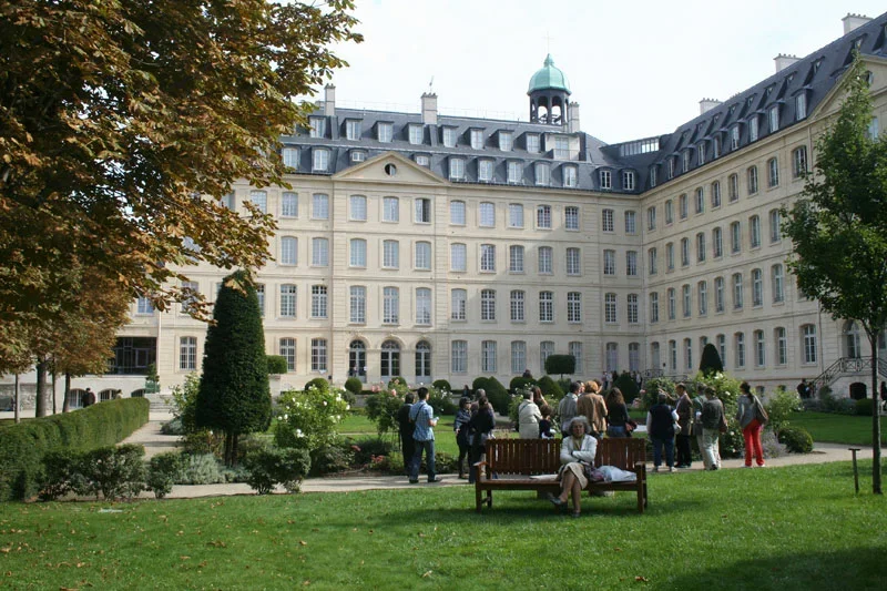Un grand bâtiment de style classique avec de nombreuses fenêtres, entouré d'un jardin bien entretenu avec des arbres et des buissons, où plusieurs personnes se rassemblent.