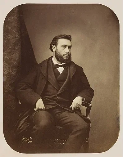 Portrait en noir et blanc d'un homme assis dans un fauteuil, vêtu de vêtements anciens, avec une barbe et des cheveux coiffés, dans un style du XIXe siècle.