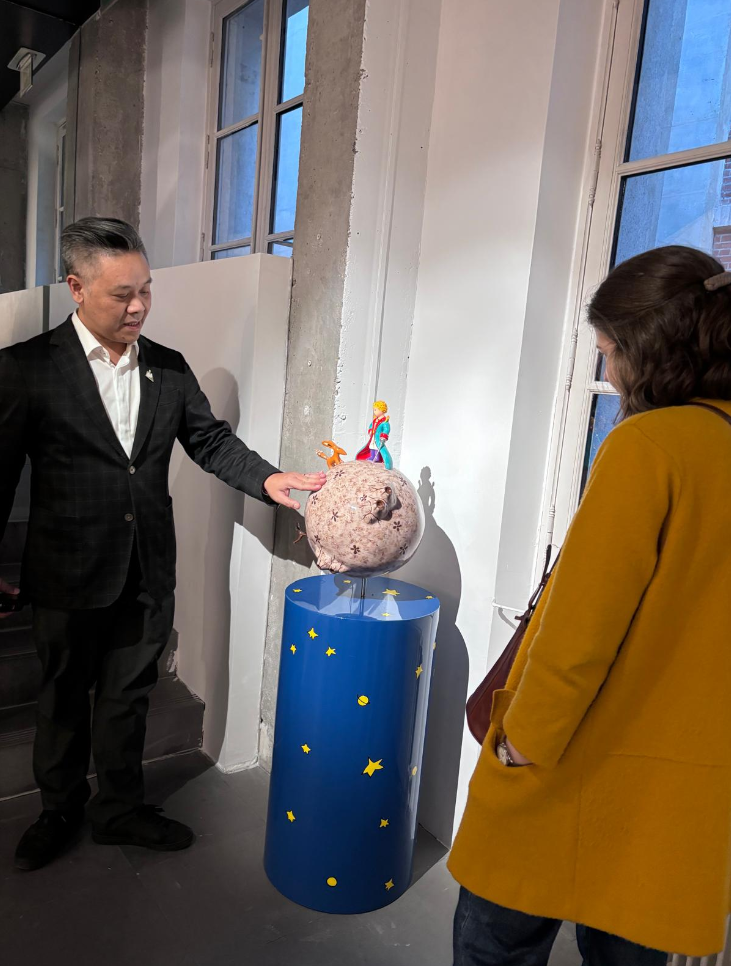 Deux personnes regardent une sculpture représentant une planète avec des personnages attachés, exposée sur une colonne bleu décorée d'étoiles et de planètes jaunes, dans une galerie d'art.