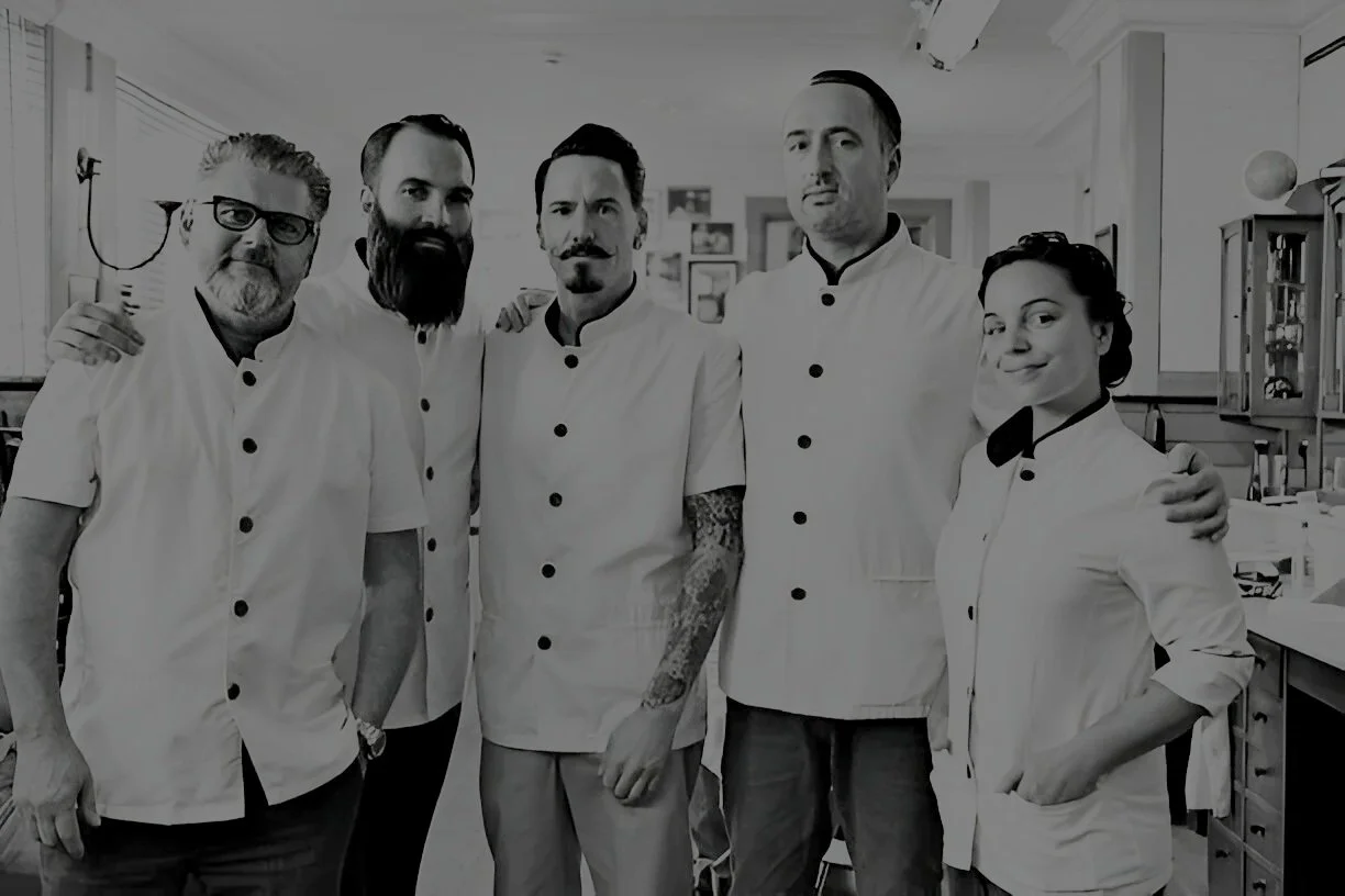 Group of five chefs standing together in a professional kitchen, wearing white chef uniforms and smiling at the camera.