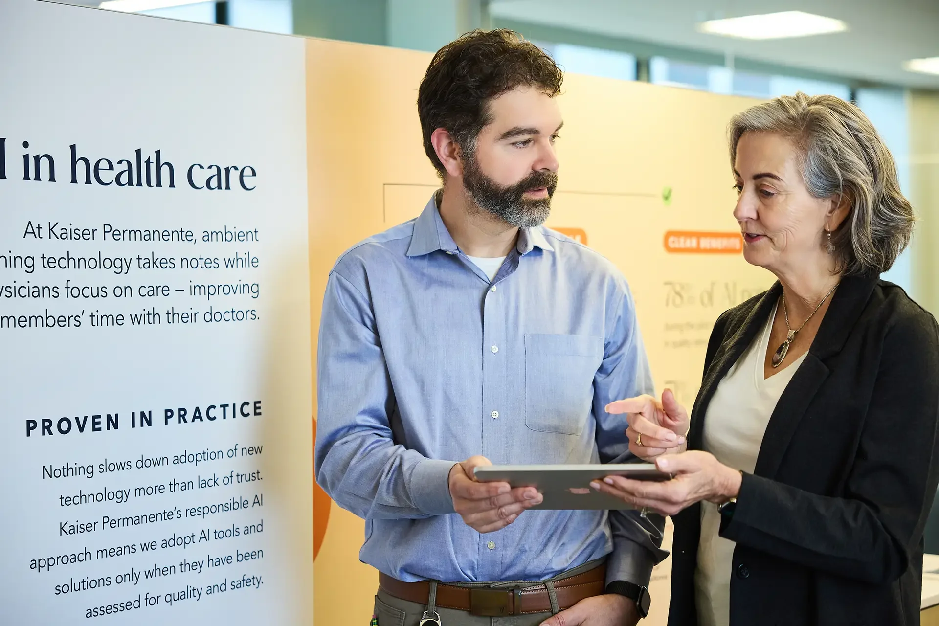 Two professionals, a man and a woman, engaged in a serious conversation at a health care conference. Behind them is a poster about health care technology at Kaiser Permanente.