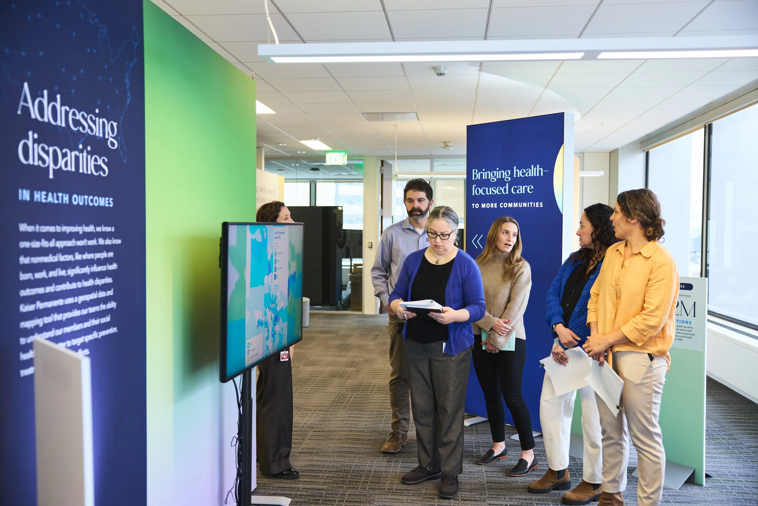 A group of five people standing in a hallway near exhibition stands with informational signs about health outcomes and communities. They are engaged and discussing, with some holding papers or notebooks.