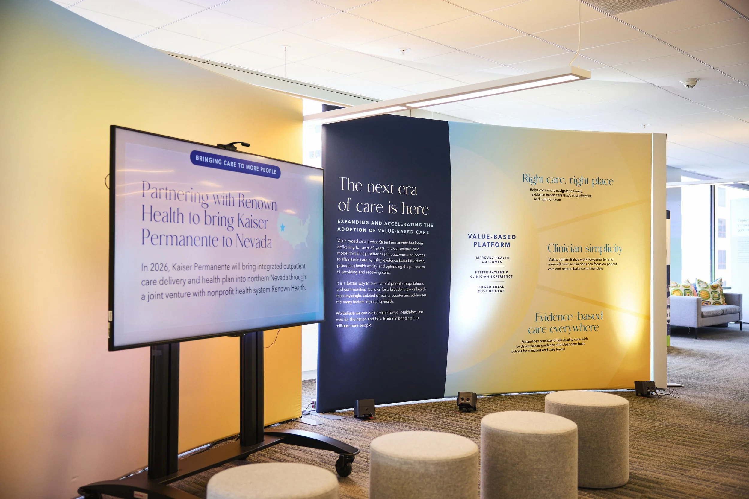 Indoor conference or seminar display with large navy and yellow informational panels and a digital screen, set in a room with carpeted floor and upholstered round stools.