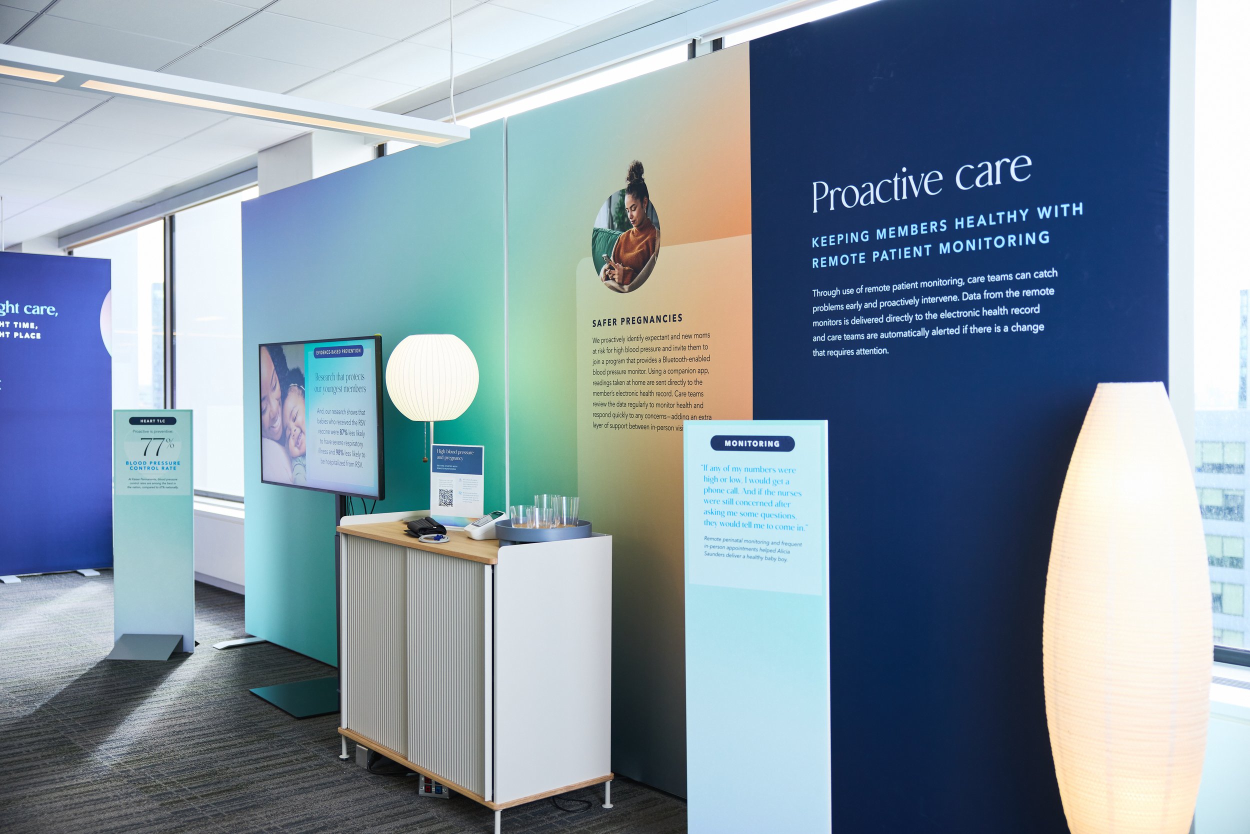 An exhibition booth displays information about proactive healthcare and remote patient monitoring, featuring a large blue and green wall with text, a monitor, a white curtain lamp, and a small table with phones and a remote control.