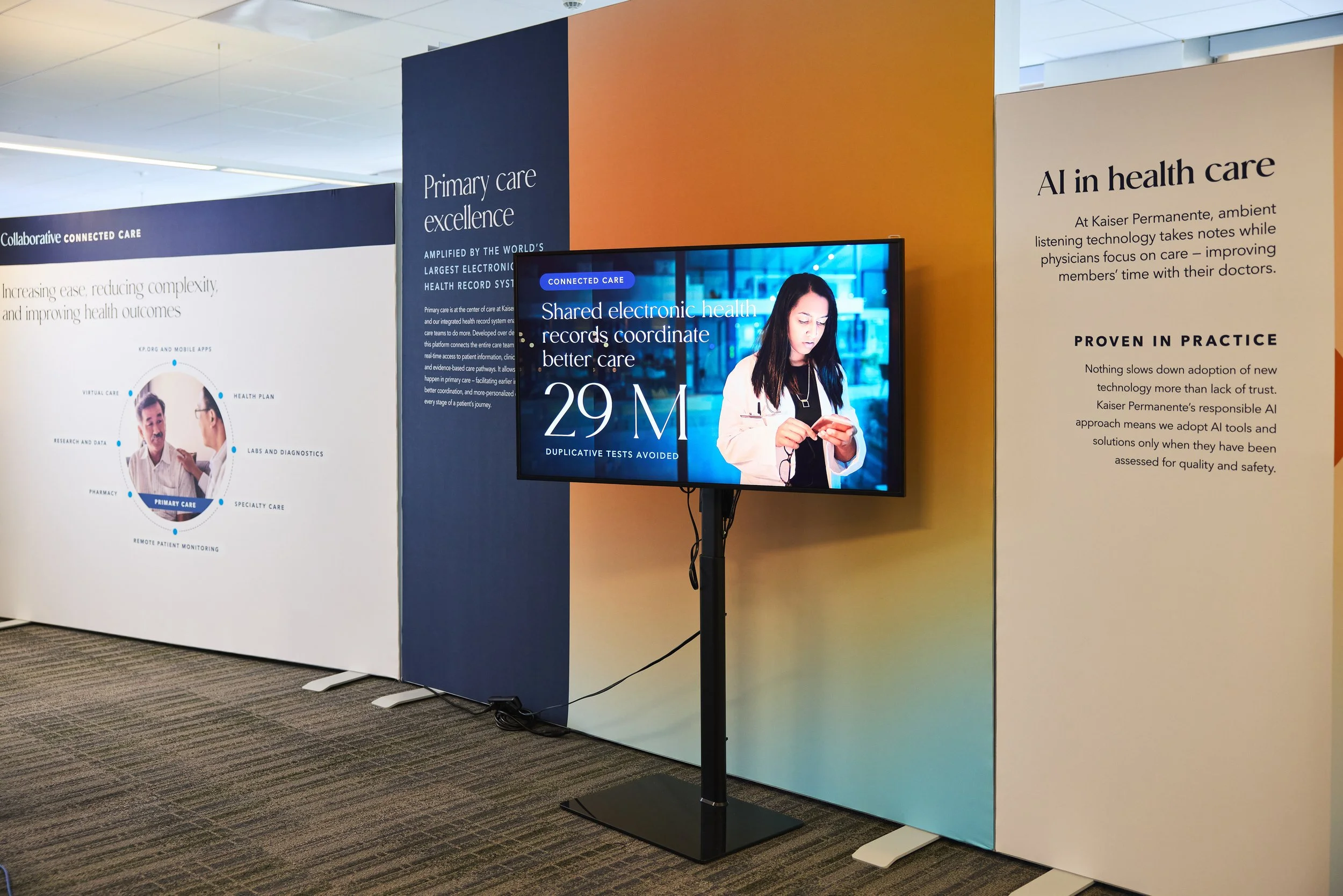 Exhibit booth with displays about healthcare technology at a conference, featuring a large screen showing statistics and text about electronic health records, AI in healthcare, and primary care excellence.