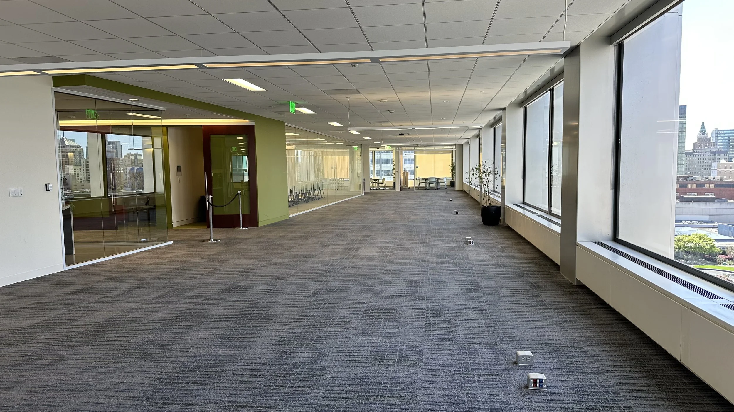 Empty office corridor with large windows, green and beige walls, glass-walled conference room, and cityscape view.