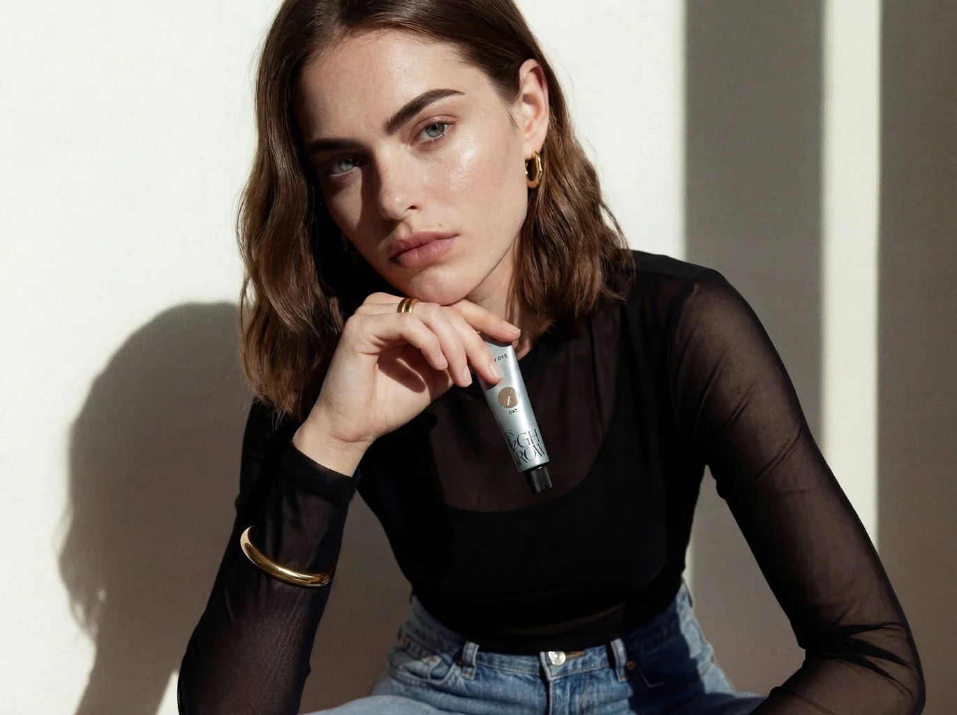 Young woman with wavy brown hair and light makeup, wearing a black sheer long-sleeve top and jeans, holding a small tube of skincare product near her face, posing indoors with a minimalist background.