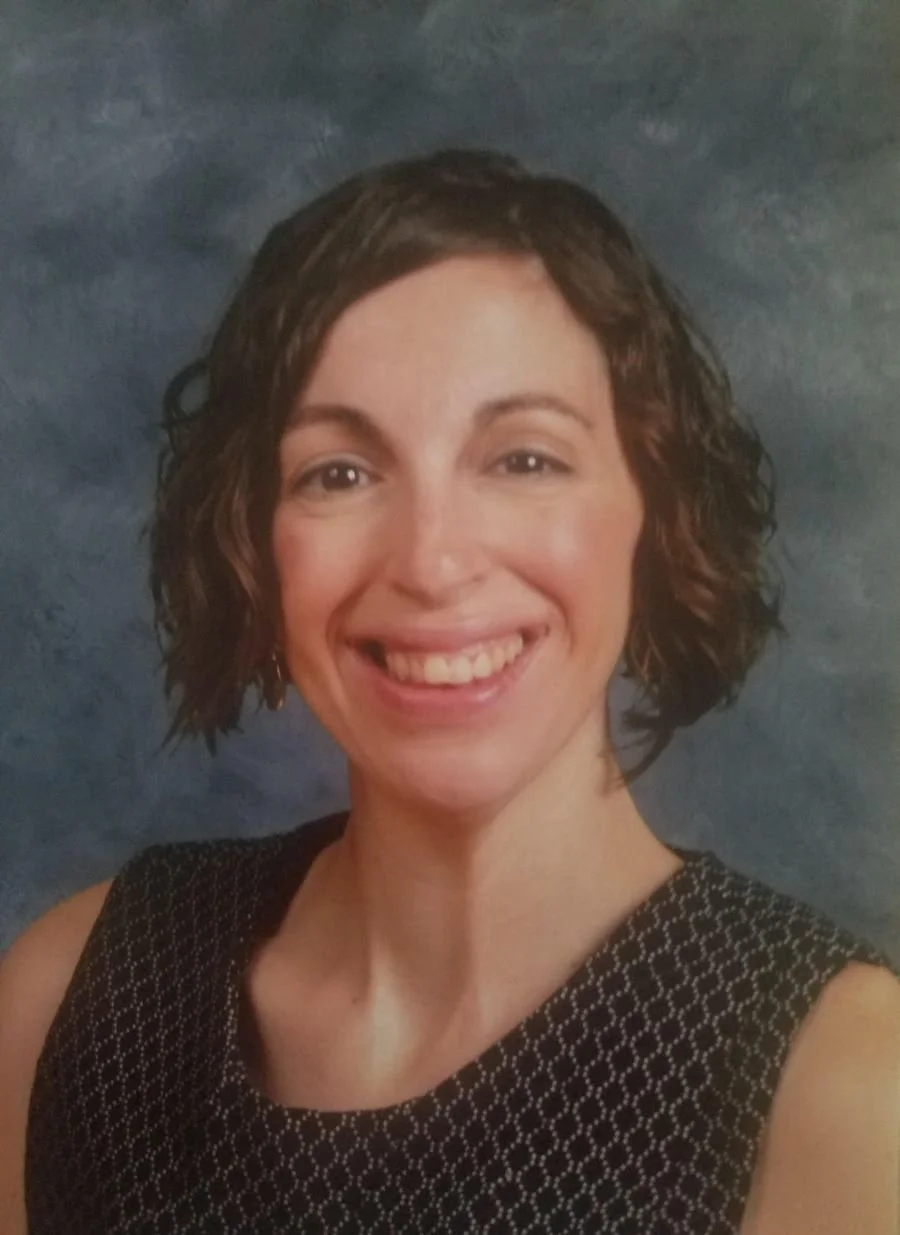 A woman with short curly brown hair smiling against a blue textured background.