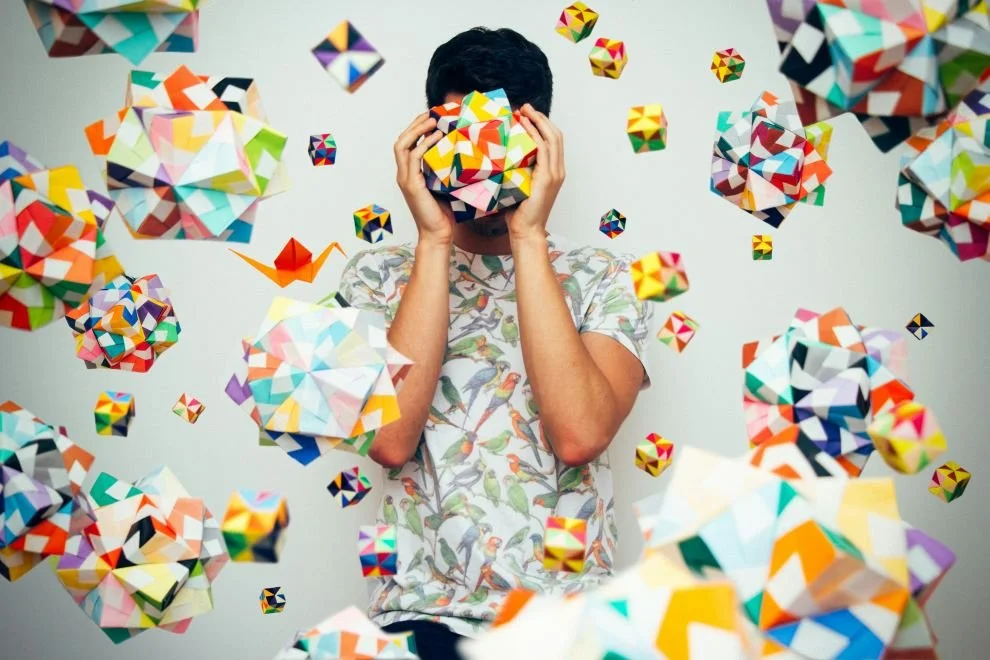 Person holding a colorful geometric object in front of face surrounded by floating similar geometric shapes.