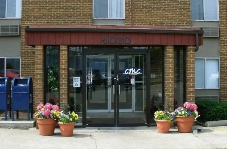 Apartment building entrance with glass doors, potted pink and white flowers, and mailboxes to the left.