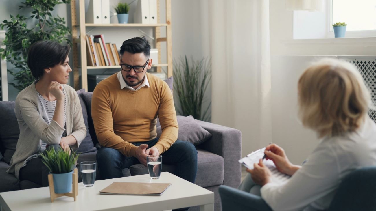 A man and woman sitting on a sofa in a therapy session with a therapist in an office, appearing upset and listening thoughtfully.
