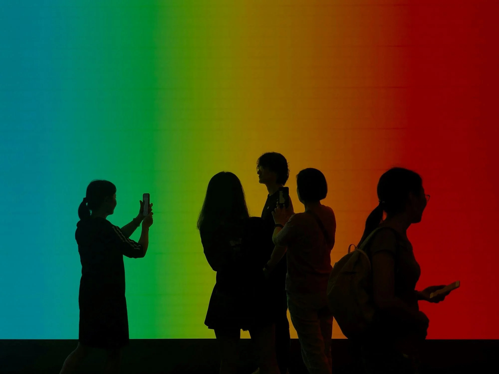 Silhouettes of people standing in front of a colorful rainbow background, some taking photos or looking at their phones.