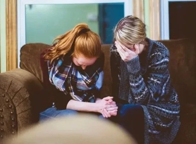 Two young women sitting on a brown couch with distressed expressions, holding hands, in a room with green and blue walls.