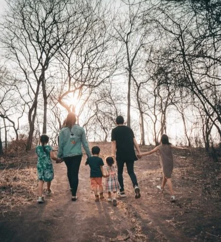 A group of five children and two adults holding hands walk up a dirt path through a leafless forest at sunset.
