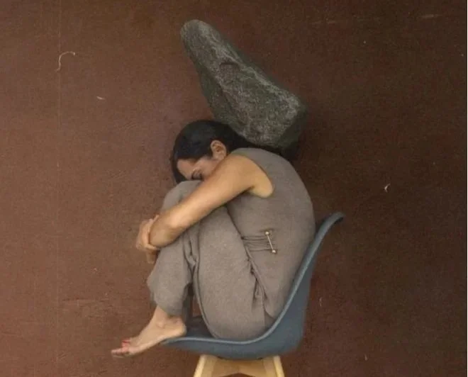 A woman sitting in a gray chair with her body curled up and her head resting on her knees; a large rock is positioned behind her head, creating the illusion of a burden trapping her