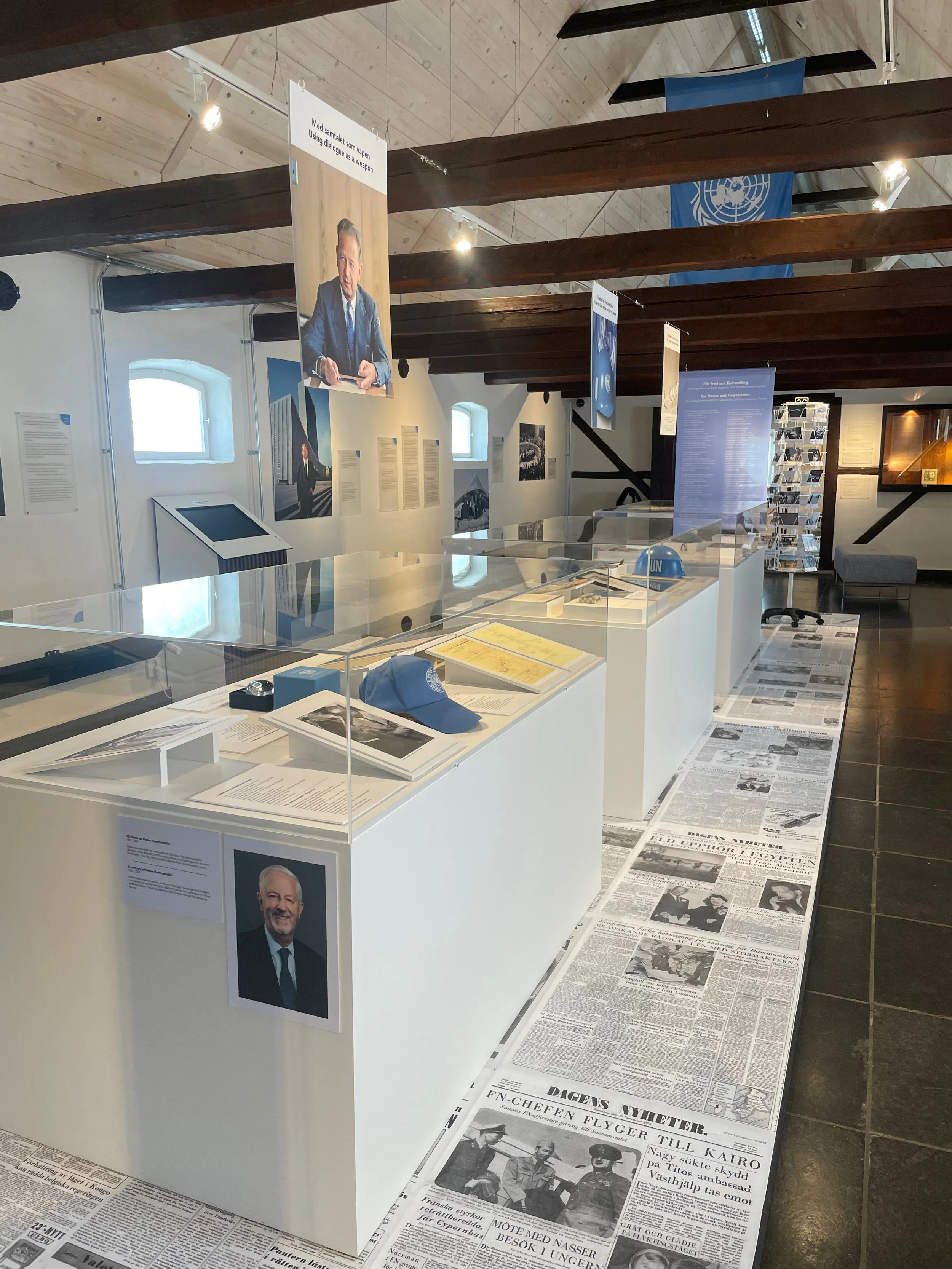 Utställning med tre vita podier med föremål och arkivmaterial. Golv av tidningsartiklar. Föremål som FN-keps och hjälm på podierna.