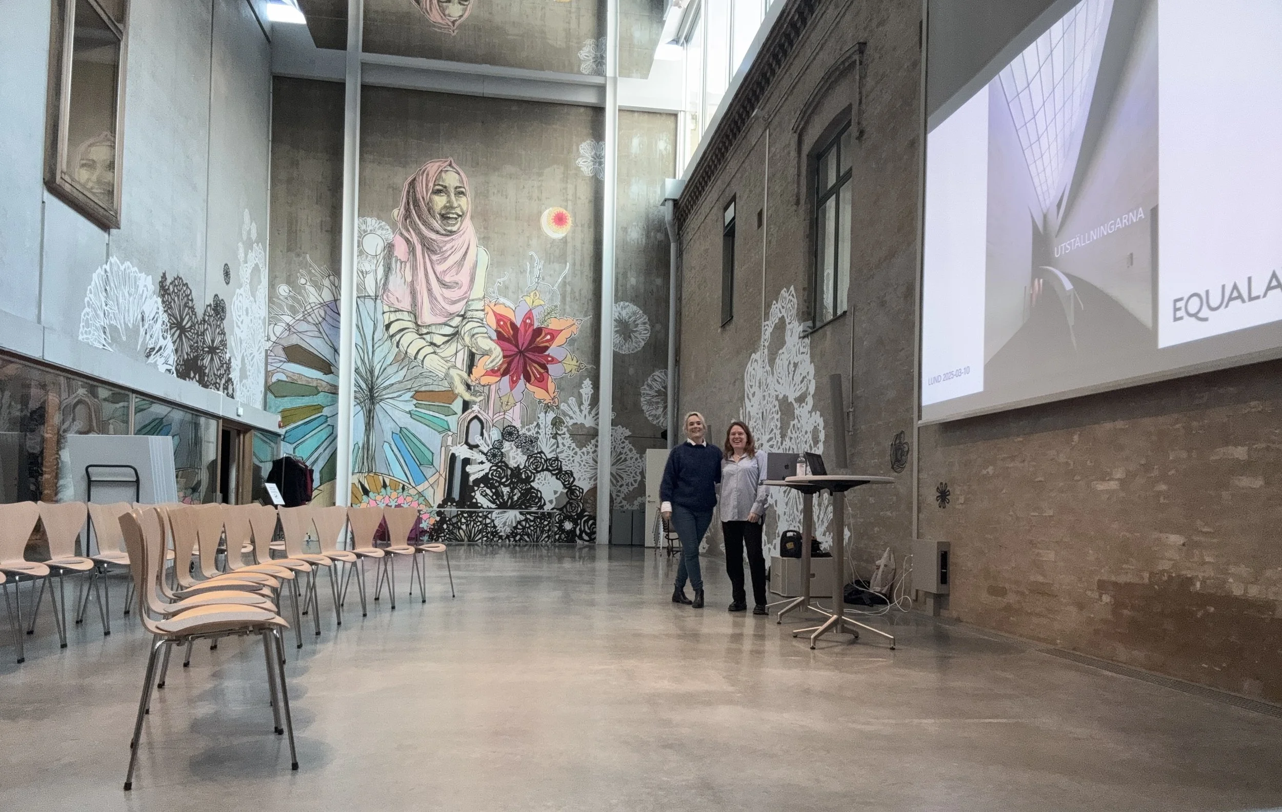 Samlingsrum på Skissernas museum med muralmålning i bakgrunden. Vid ena sidan står Sofie och kollega Johanna Lundin.