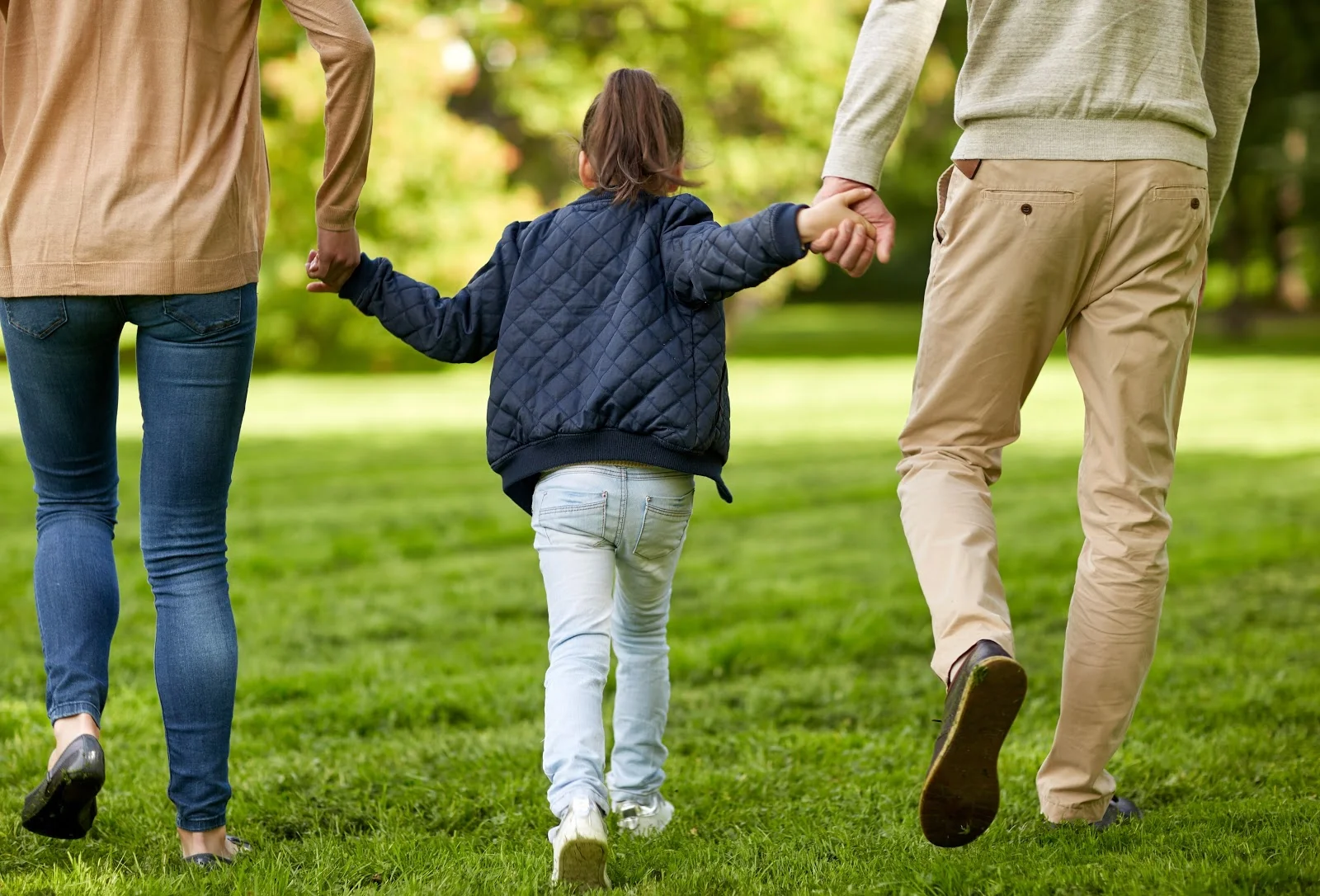Little girl with one hand held by each parent walking through the grass
