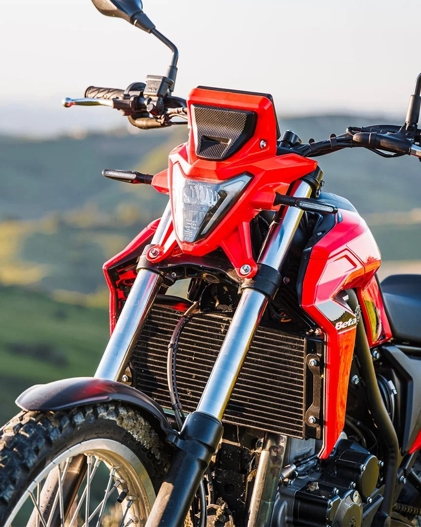 A red off-road motorcycle with a high front fender, suspension, and radiator, set against a blurred outdoor landscape.