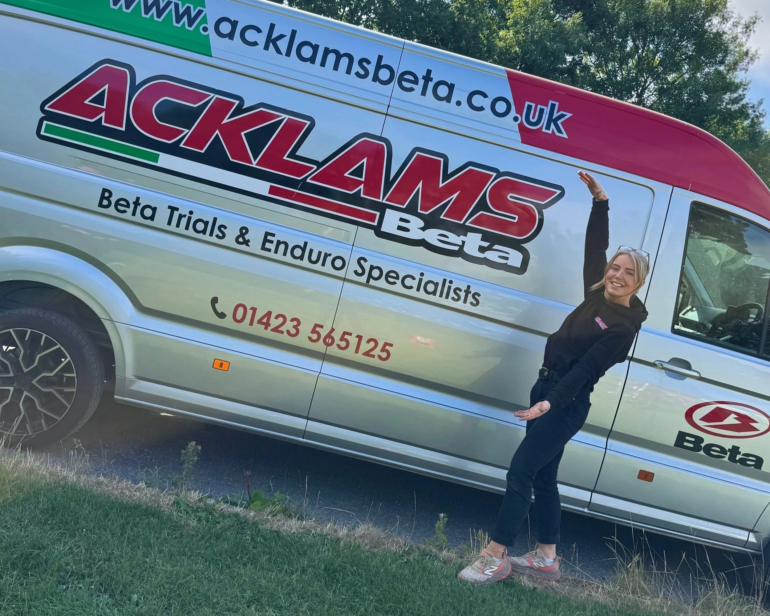 Morgan standing next to a promotional vehicle for ACKLAMS Beta, a company specializing in beta trials and enduro racing, outdoors with trees in the background.