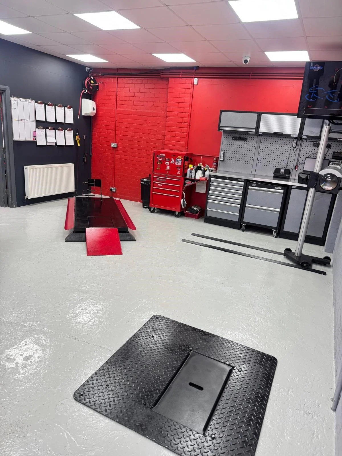 A professional automotive workshop with red and gray tool cabinets, a red rolling toolbox, a workbench with tools and supplies, and a vehicle lift in the foreground.