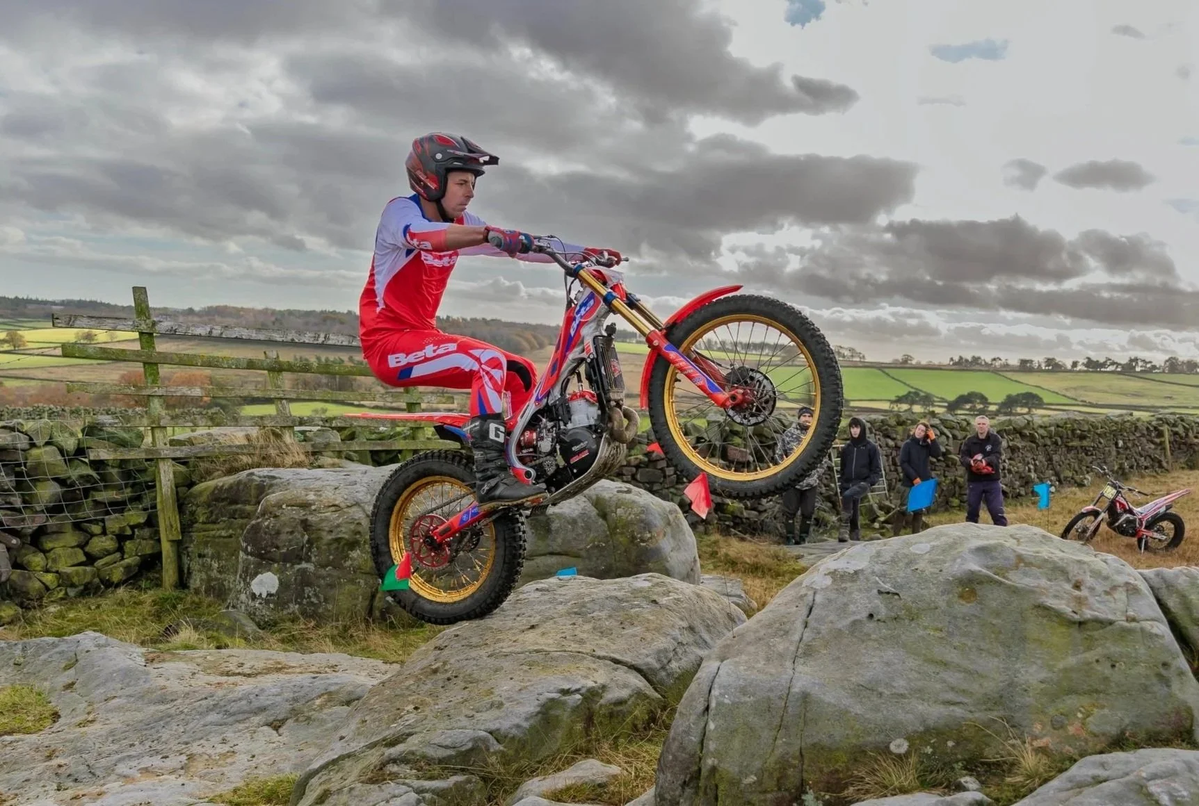 Joel Sadler, Acklams director riding a trials bike over large rocks in an outdoor rural area, with spectators watching behind a stone wall.