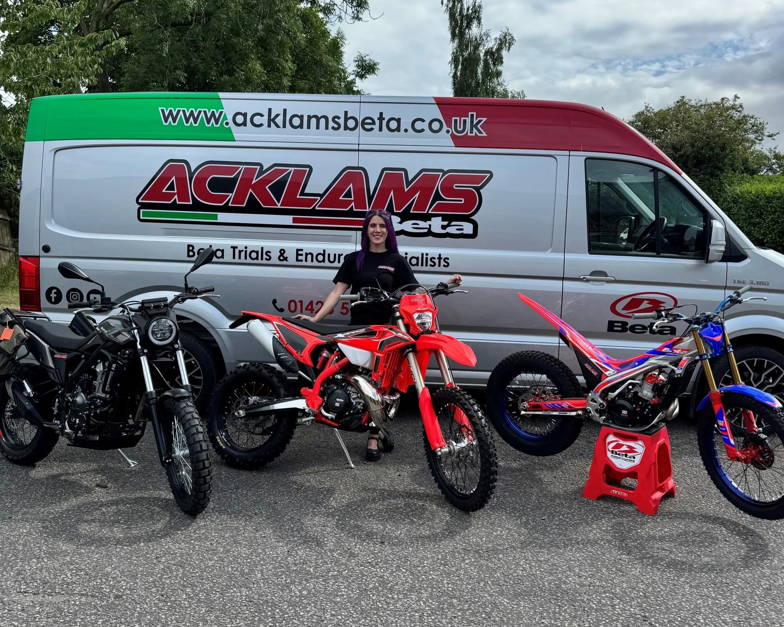 Jessica in sales and parts standing next to a red and white dirt bike and other motorcycles, in front of a large van with logos and website URL for Acklam Beta on it, in an outdoor setting with trees and a cloudy sky.
