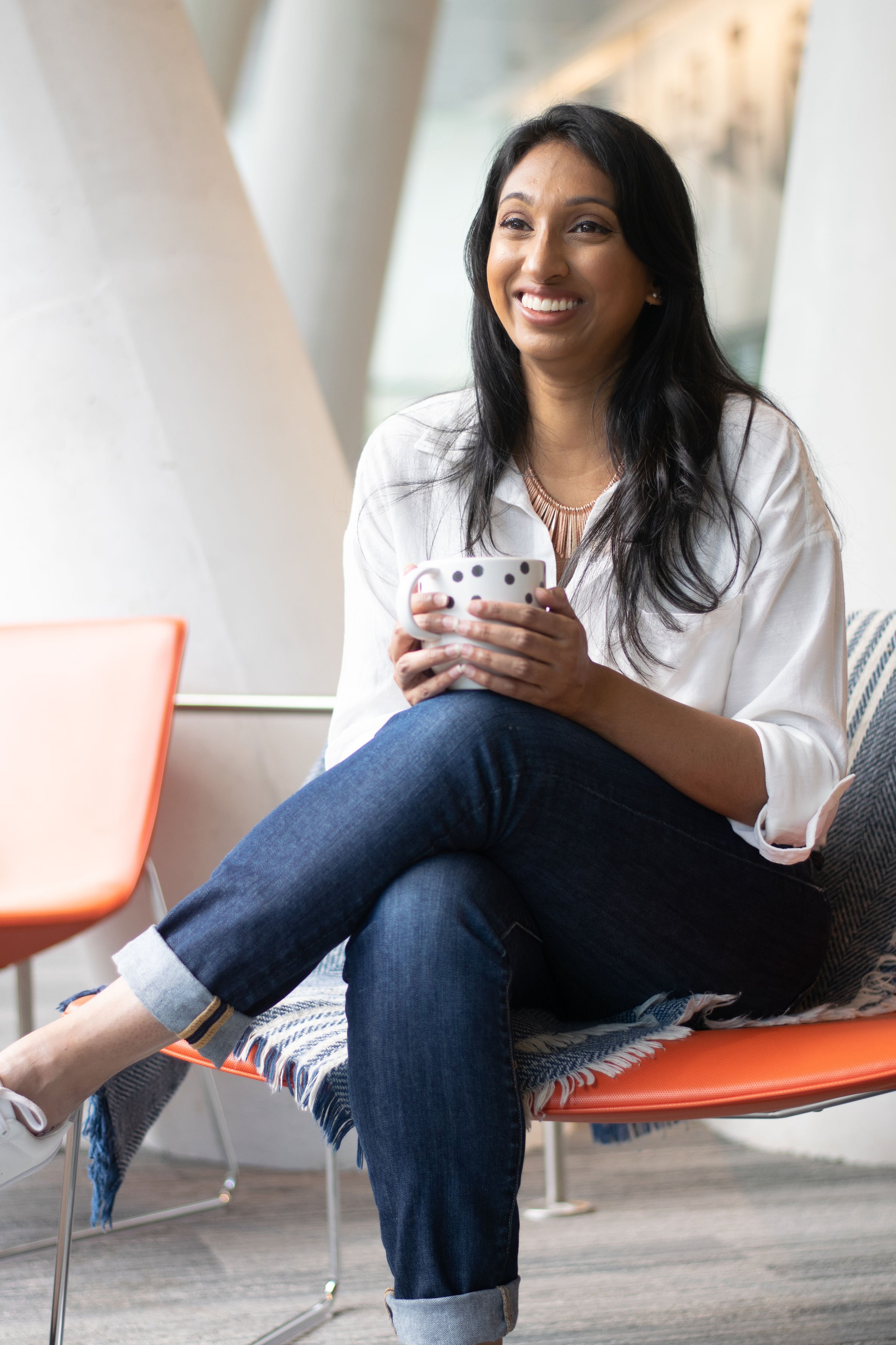 Smiling Indian woman sitting in an orange chair that is partiallly covered with a throw blanket, with one knee crossed over the other, resting a coffee cup held in both hands on the top knee.