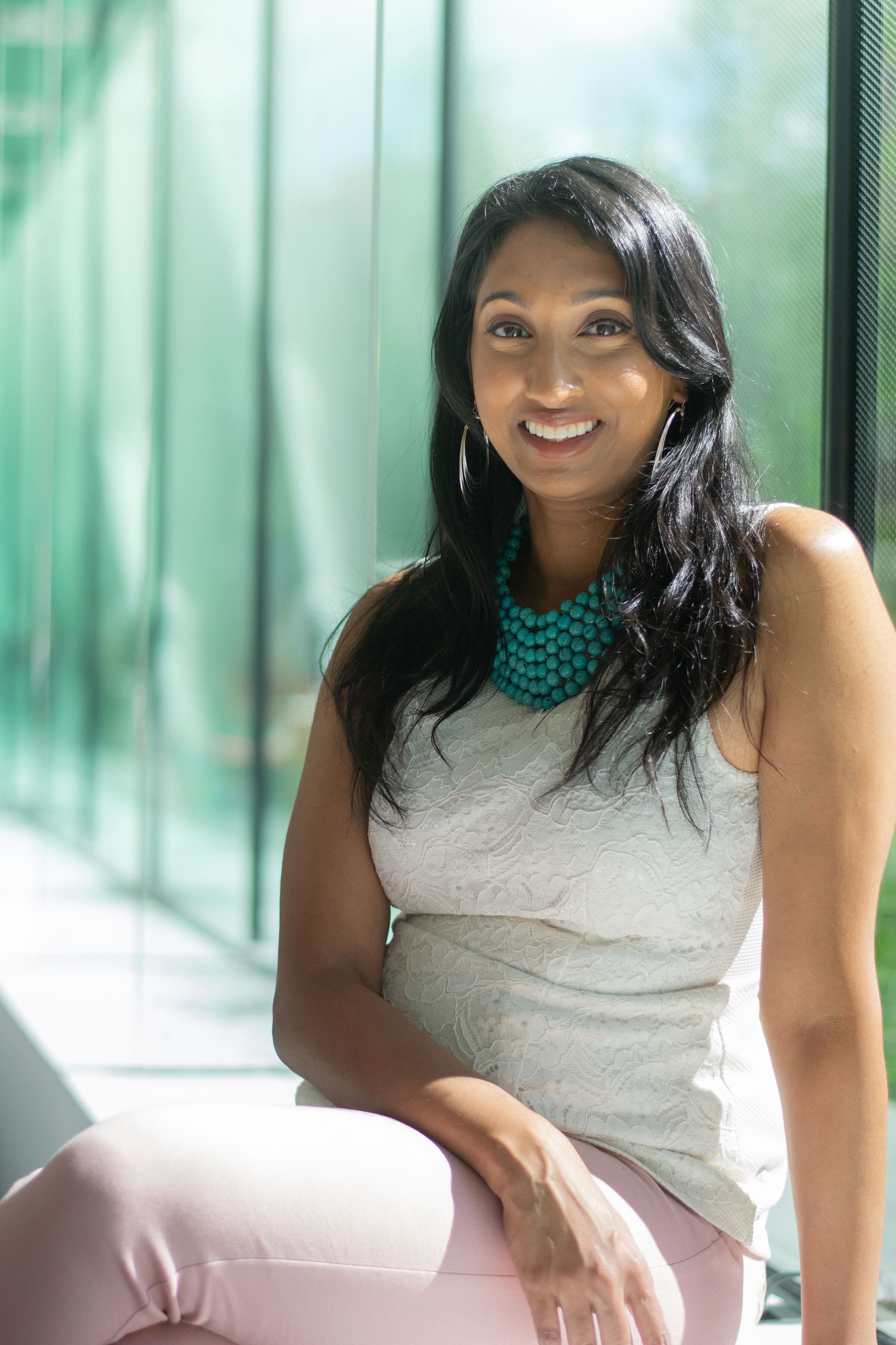 Indian woman with silver hoop earrings, aqua colored bead necklace, white top, pink pants, sitting at an angle on a small white ledge in front of green tinted glass windows