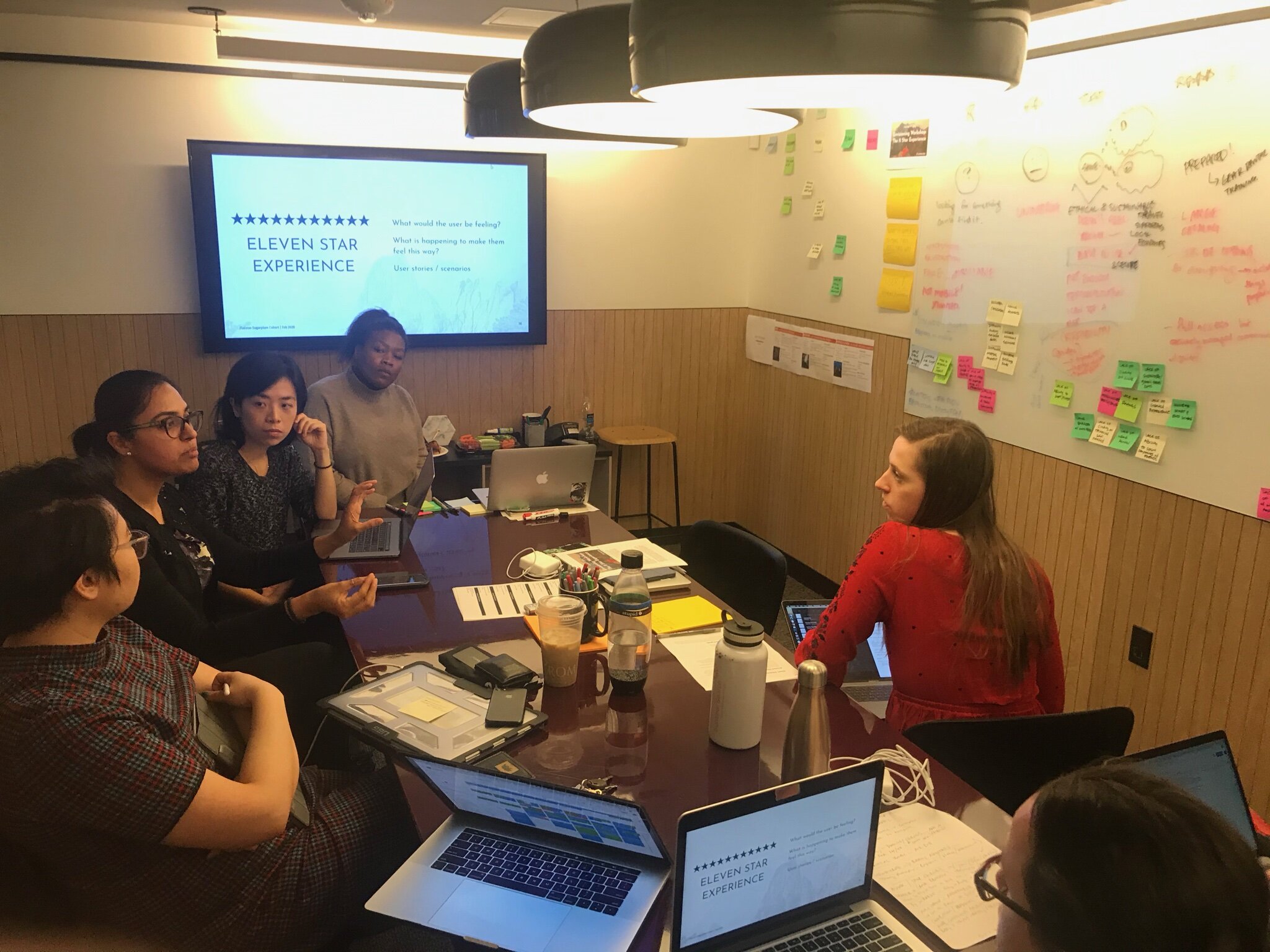 A photograph shows several people sitting around a wooden conference table in a room with wood-paneled walls, looking at a whiteboard covered in colorful sticky notes and a television screen.
