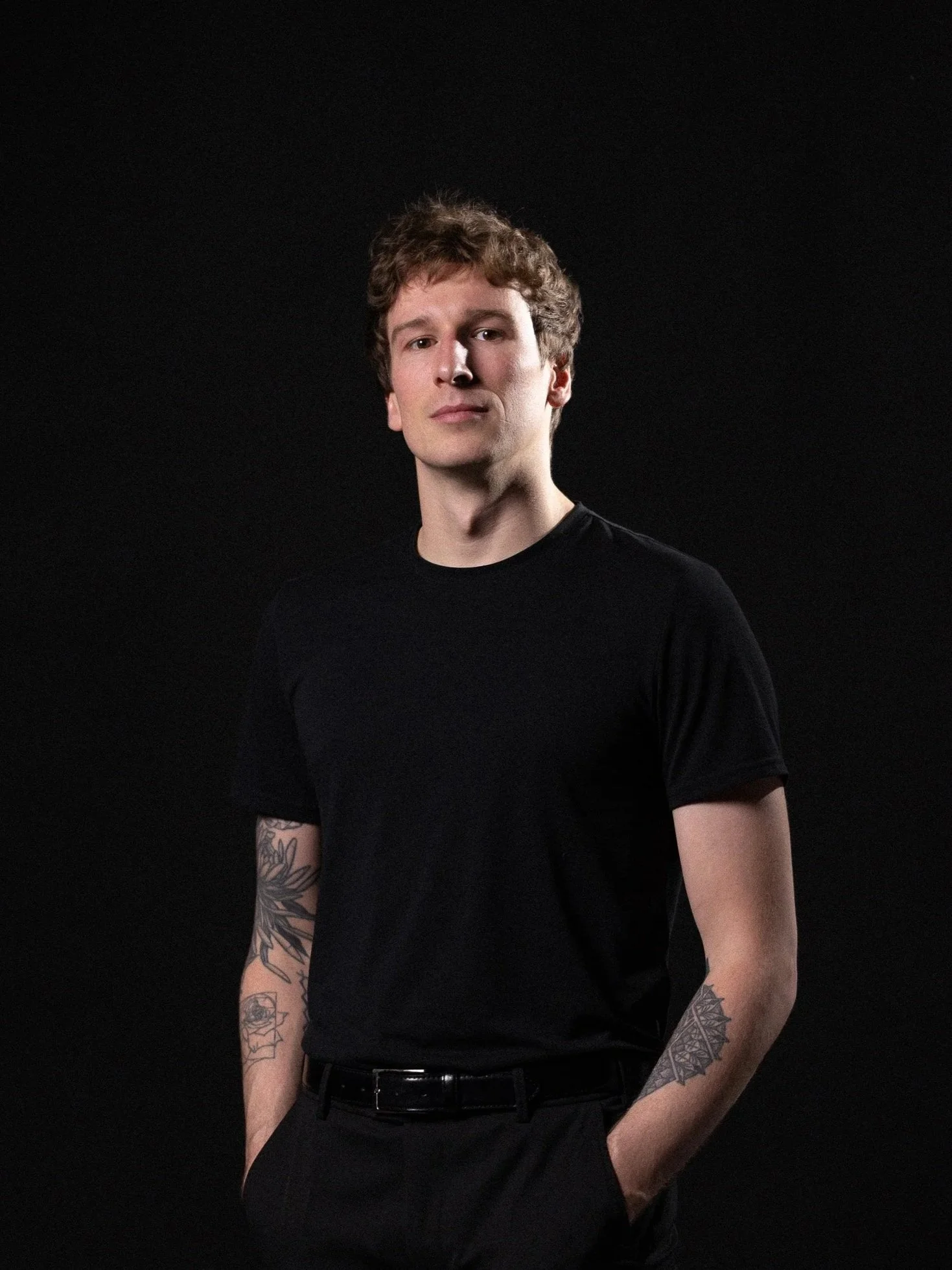 A young person with curly brown hair and light skin, wearing a black T-shirt, standing against a black background. They have tattoos on both arms and are standing with hands in pockets.