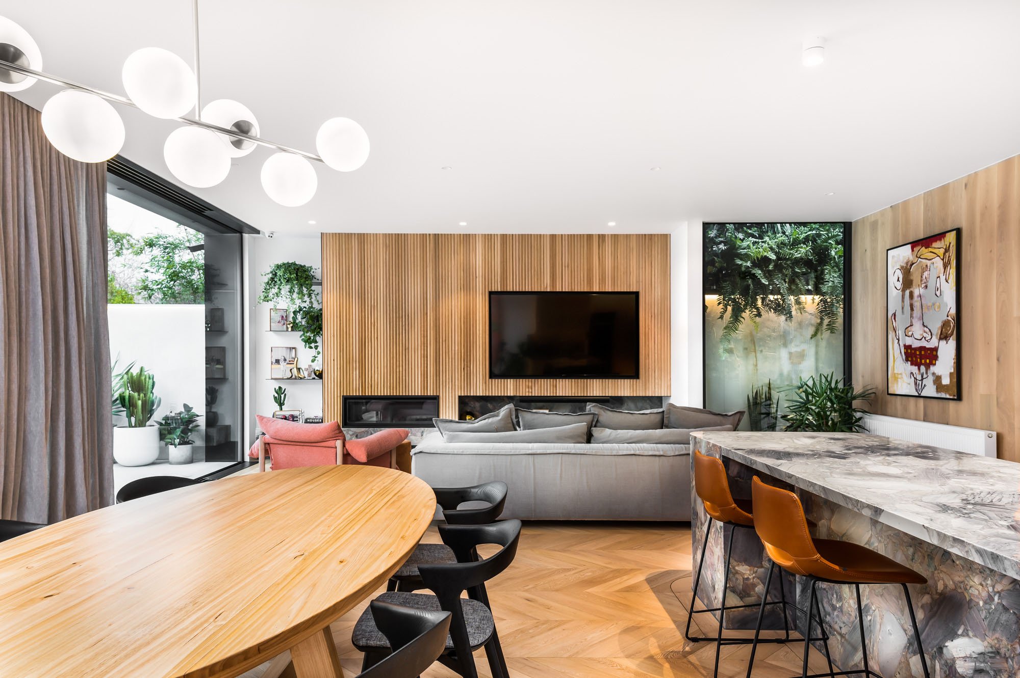 Modern living room with wood-paneled accent wall, mounted flat-screen TV, gray sofa, pink armchair, marble kitchen island with orange chairs, indoor plants, abstract artwork on wall, chandelier with globe lights, and large window with curtains.