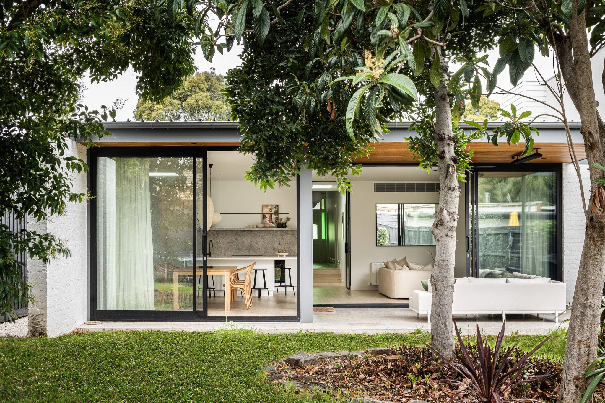 Modern house exterior with large glass sliding doors, white walls, wooden ceiling accents, and lush landscaping with trees and grass.